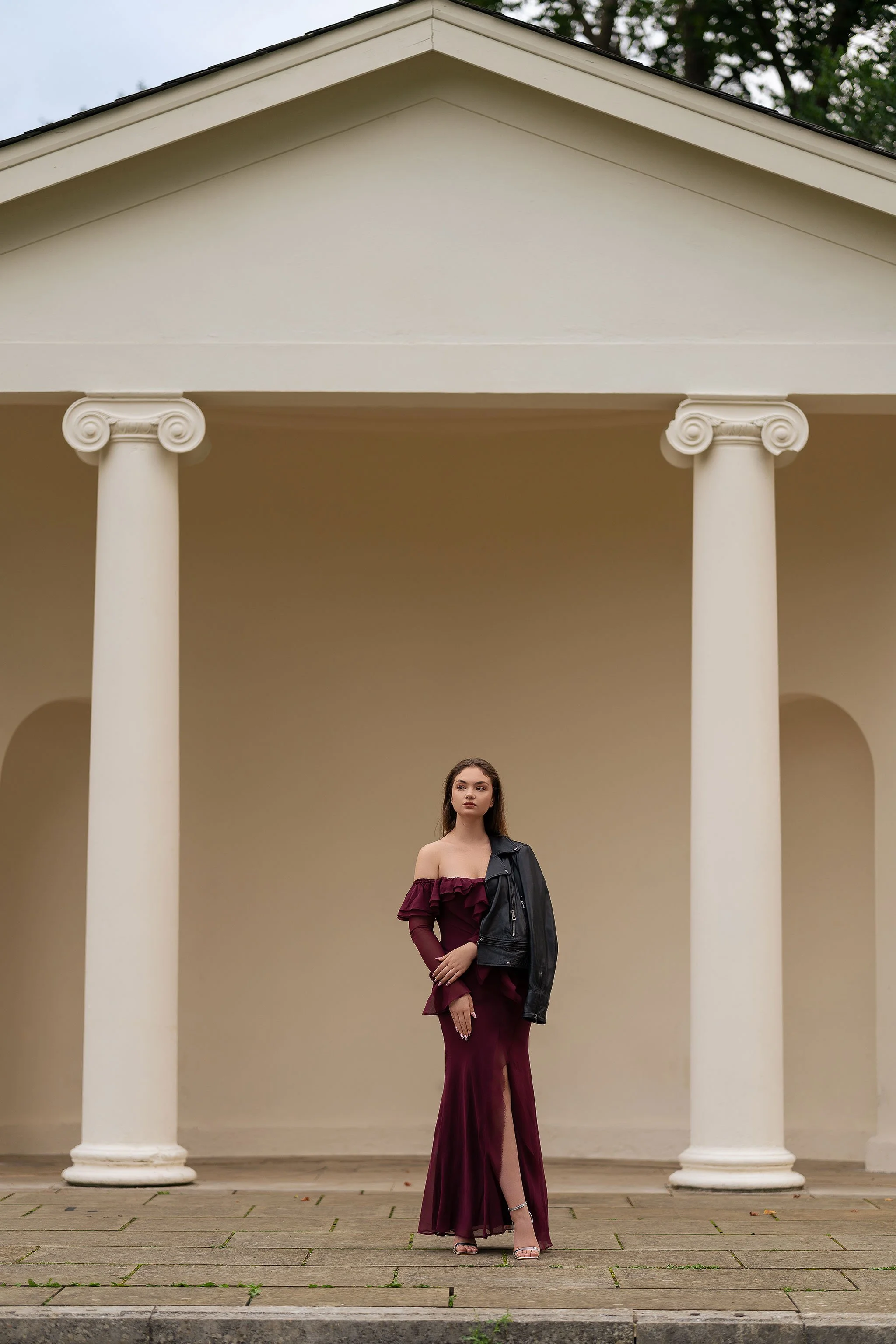 Striking outdoor fashion portrait of a woman blending elegance and edge — wearing a burgundy dress and leather jacket, framed by neoclassical architecture.