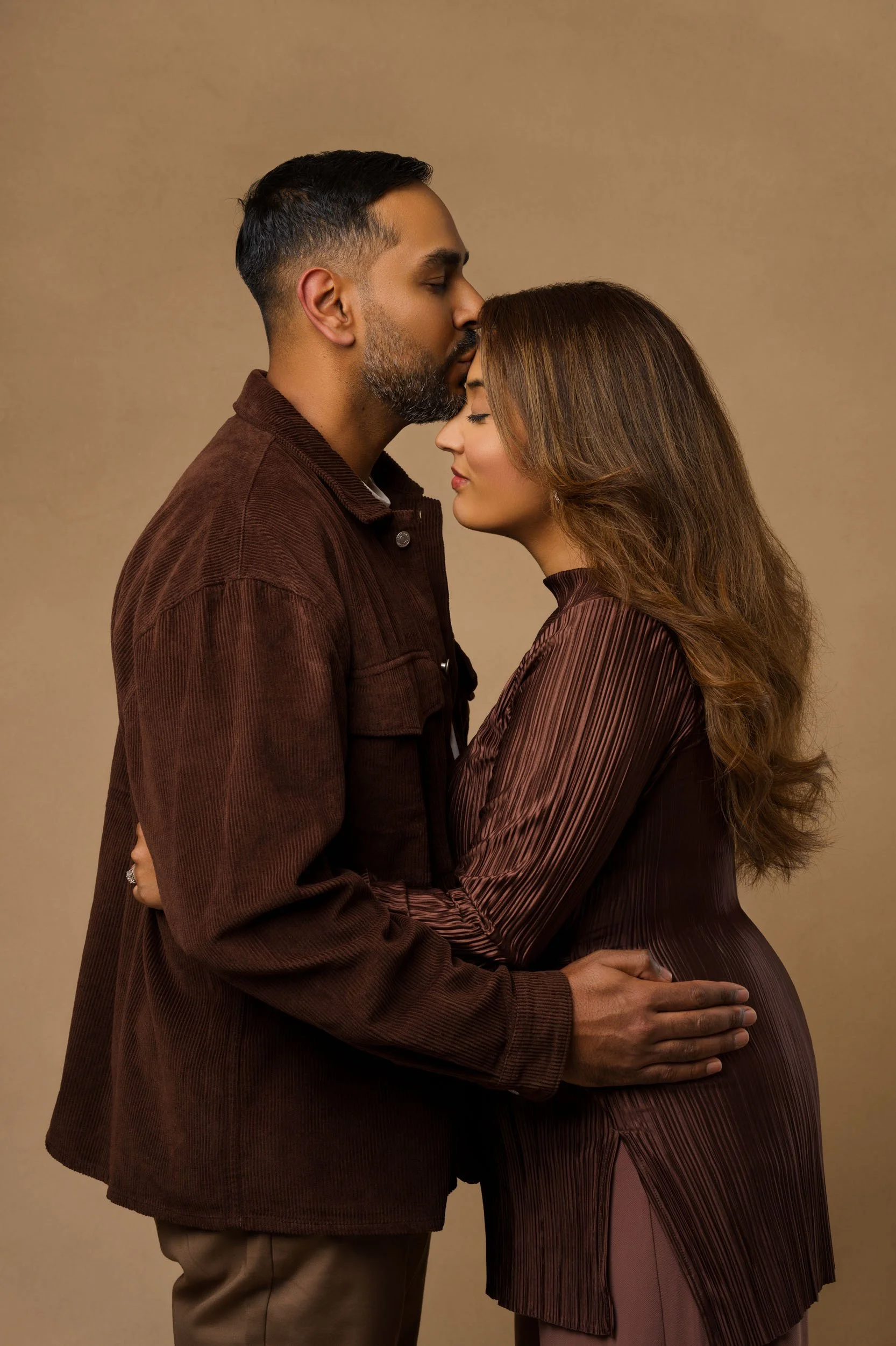Studio couple on tan backdrop, man kisses woman’s forehead while they hold each other in a gentle embrace.