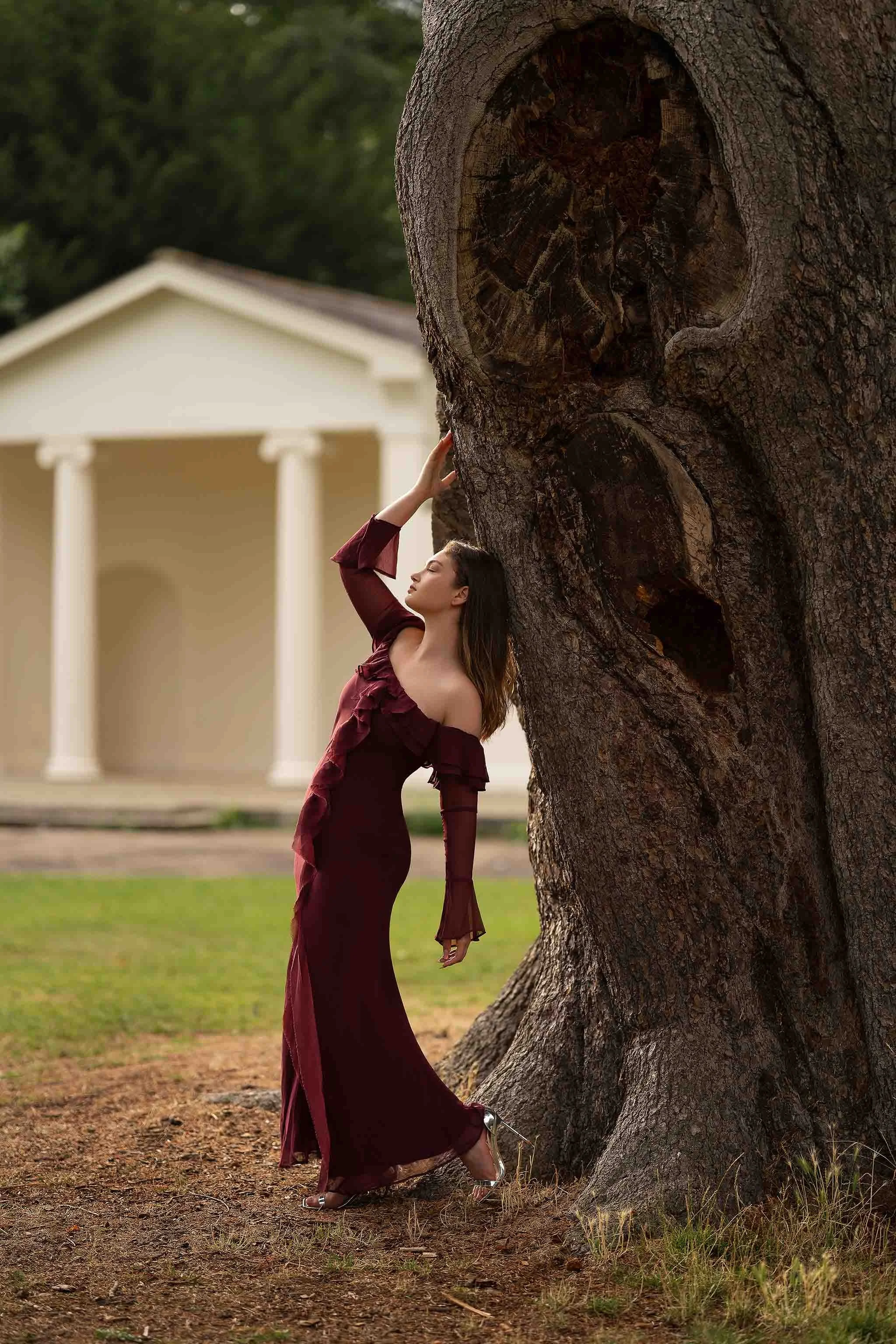 Graceful outdoor portrait featuring a woman in a burgundy gown posed beside an ancient tree—elegant, timeless, and full of character.