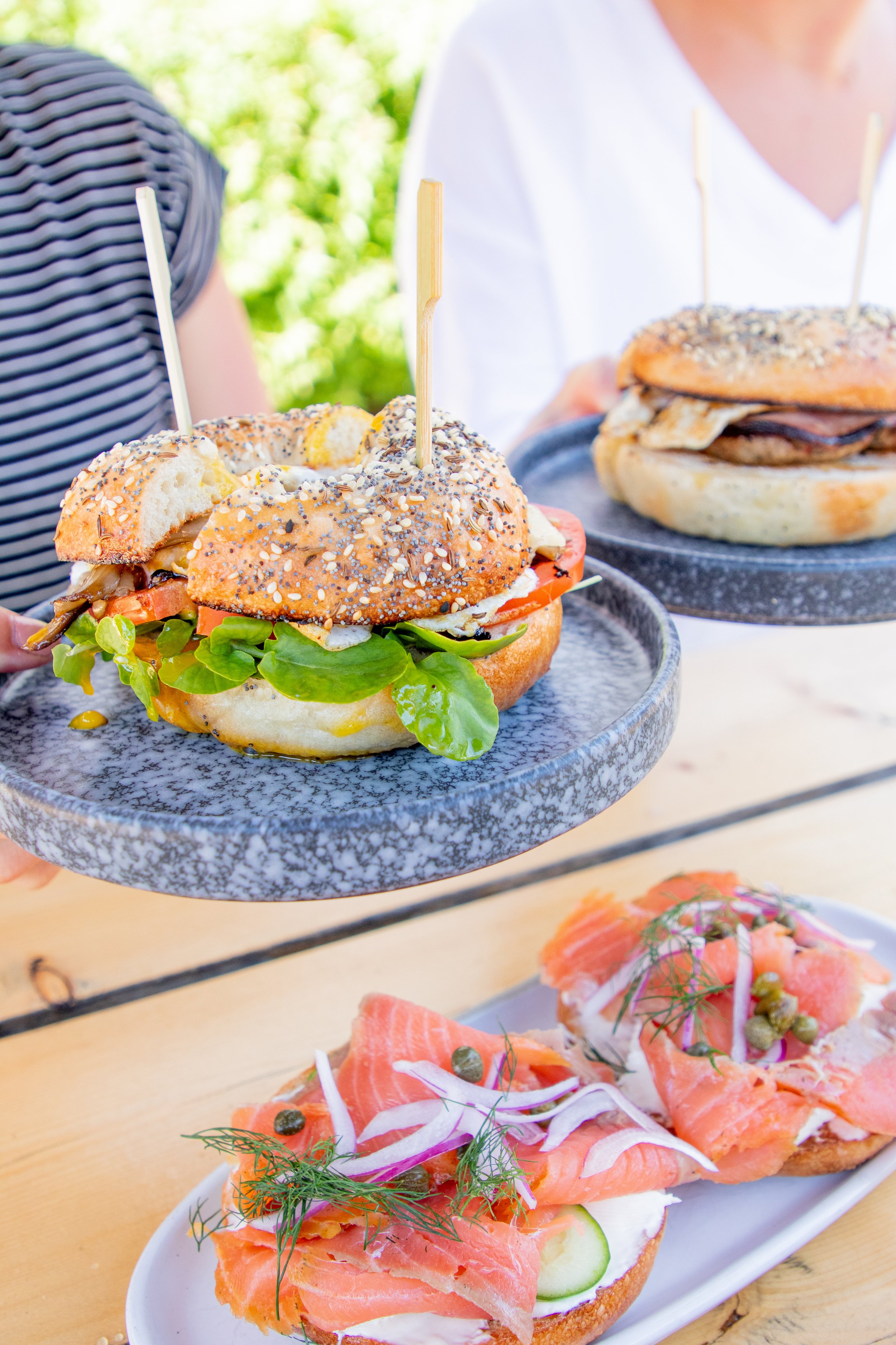 A person holding a plate with a bagel sandwich containing lettuce, tomato, and other ingredients, and a skewer topping. Another person holds a sandwich with a bagel bun, and a platter in front has smoked salmon on toasted bread with onion slices, cucumber, and herbs. The background shows greenery and a wooden table.