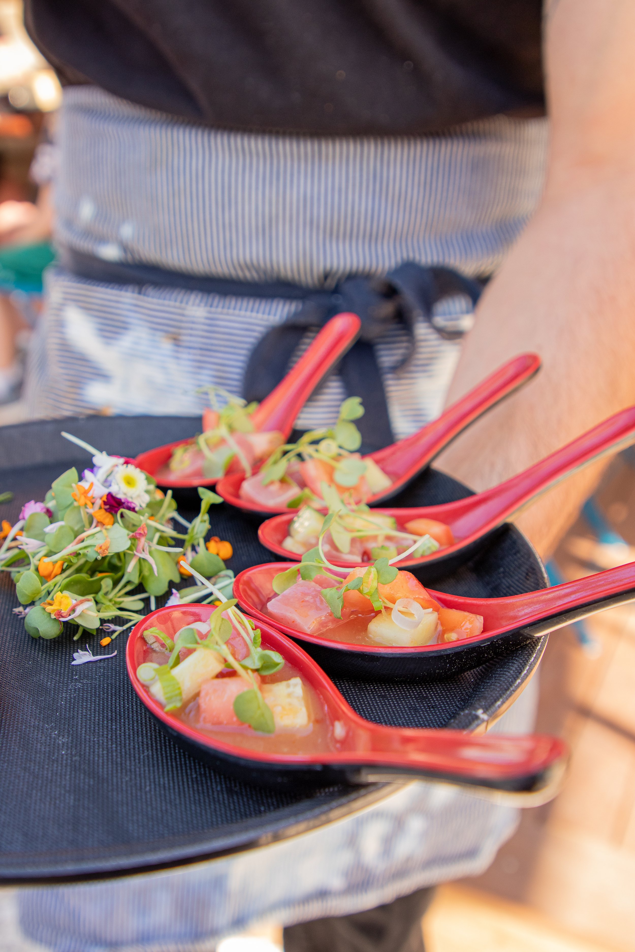A serving tray with small red bowls of colorful seafood salad and microgreens, held by a person wearing an apron.