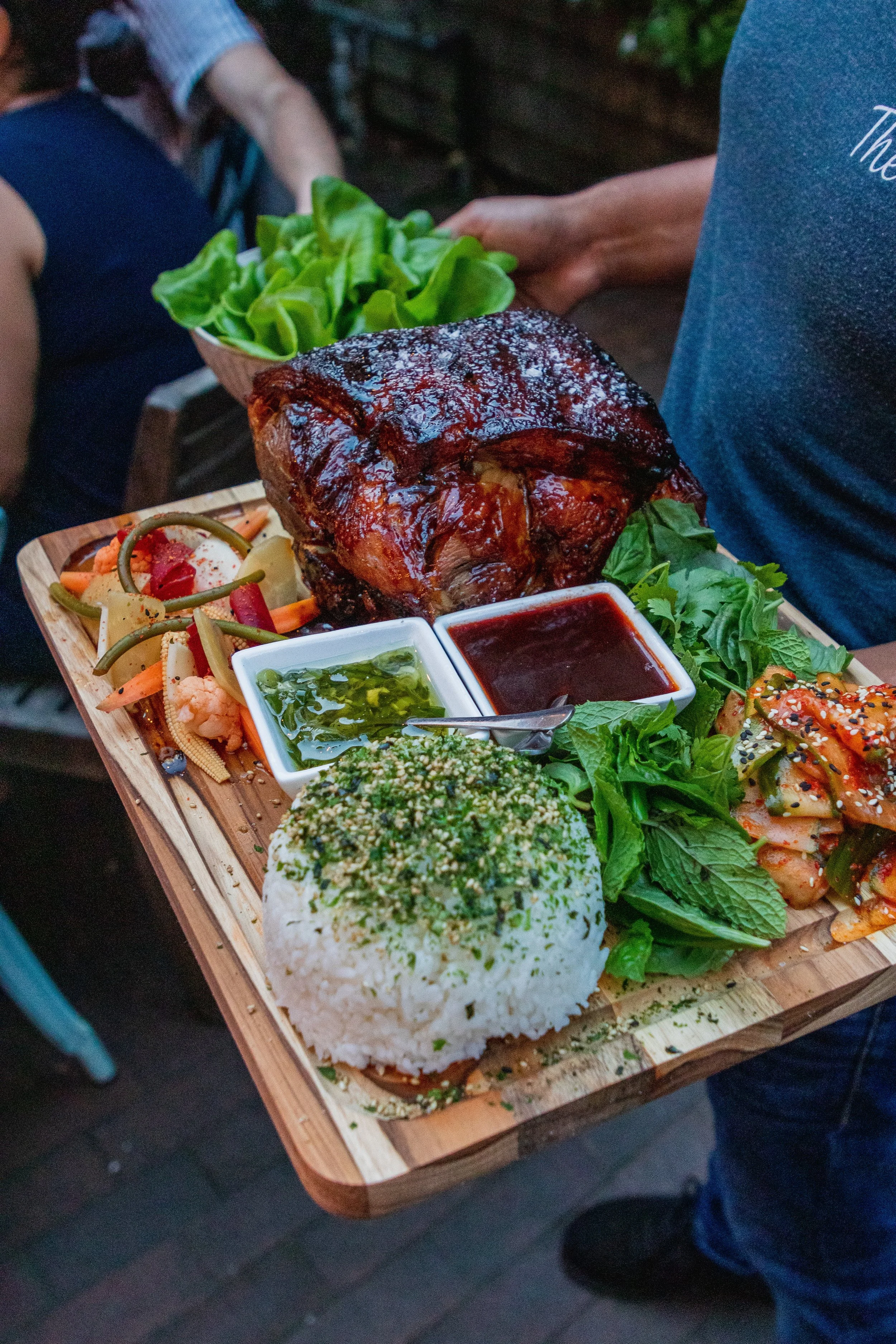Tray of Asian-inspired dishes including rice, grilled meat with sauce, fresh greens, vegetables, and condiments.