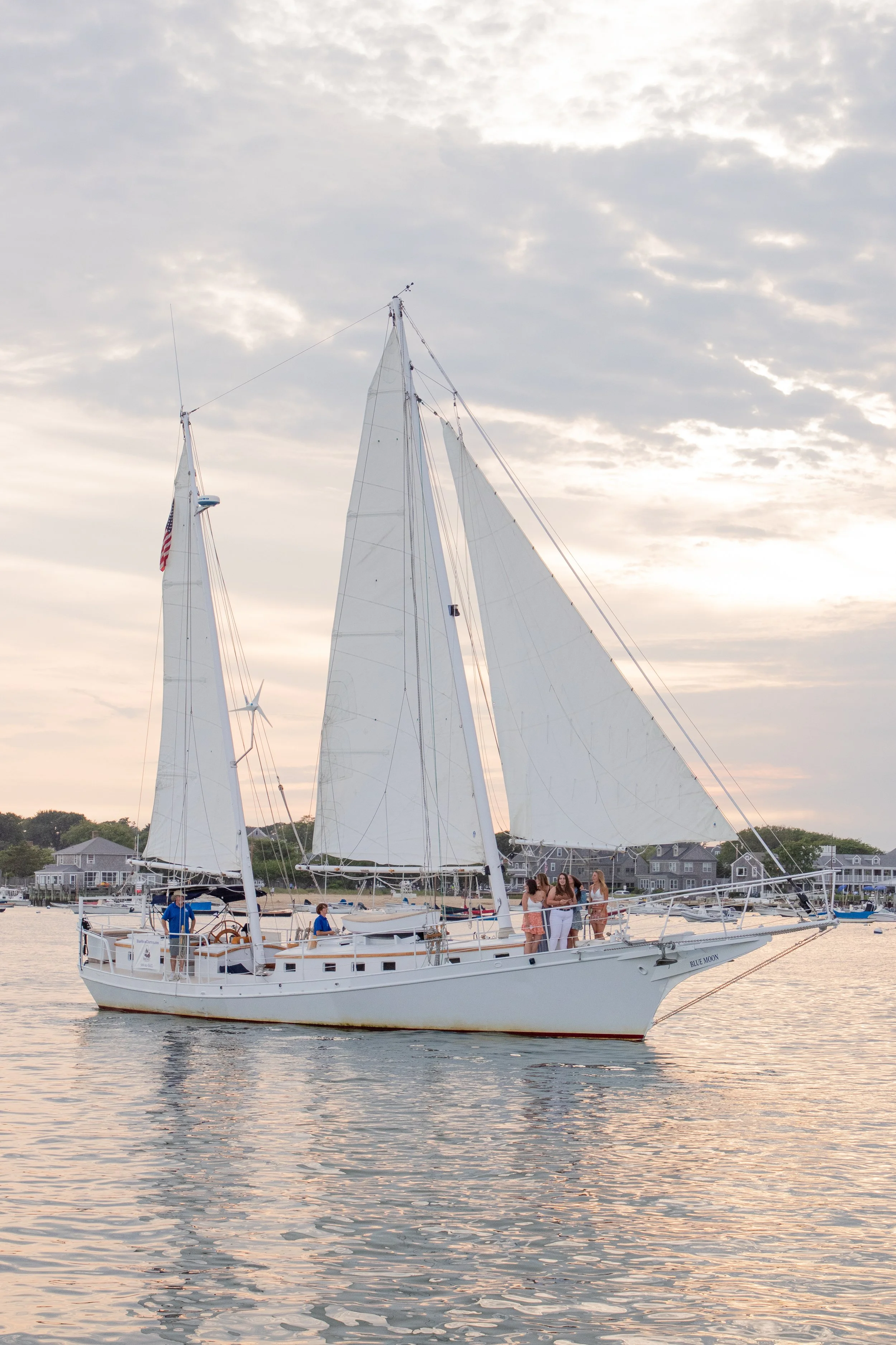 A sailboat on calm water during sunset with five people onboard, some standing and chatting, in a scenic harbor setting.