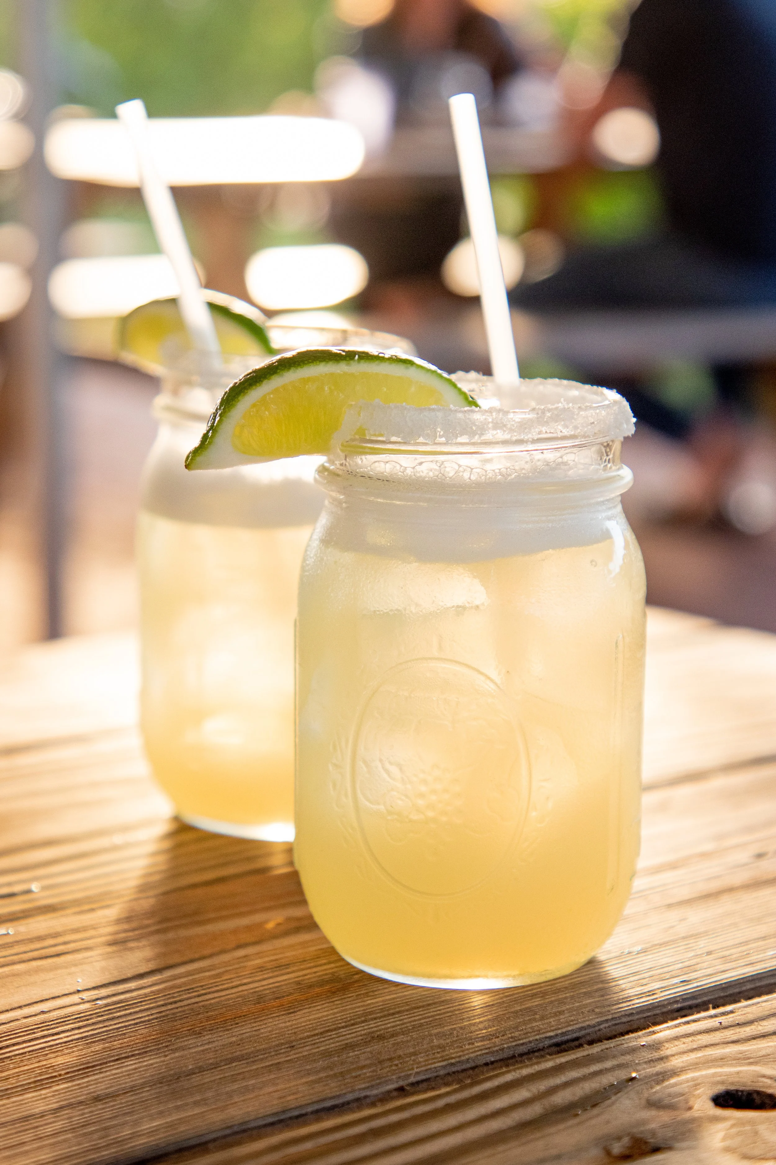 Two mason jars filled with a light yellow lemonade, each garnished with a lime wedge and topped with a salted rim, placed on a wooden table outdoors.