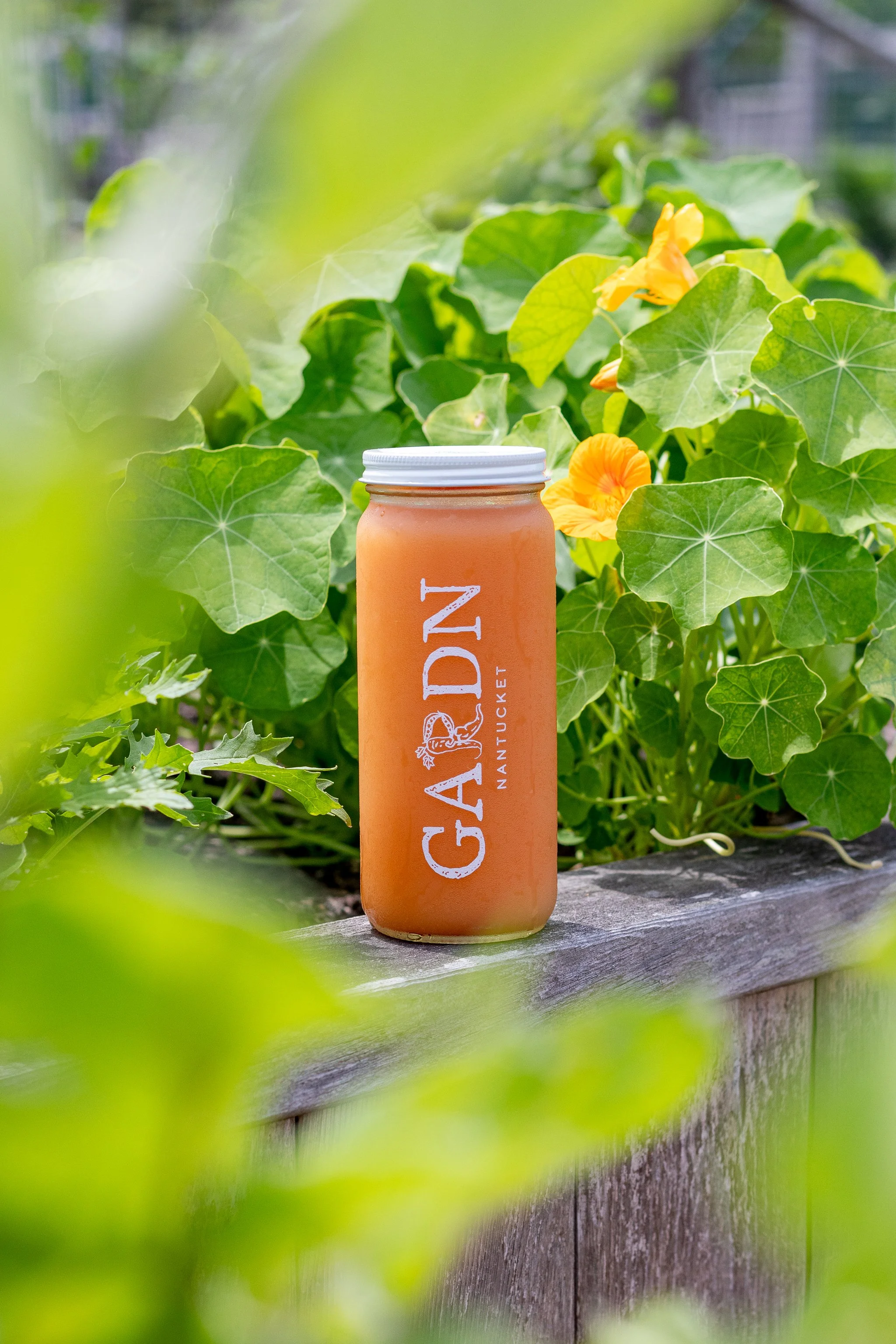 A mason jar of juice with a white lid, labeled 'GARDEN NANTUCKET,' sitting on a wooden ledge surrounded by green leaves and yellow flowers.
