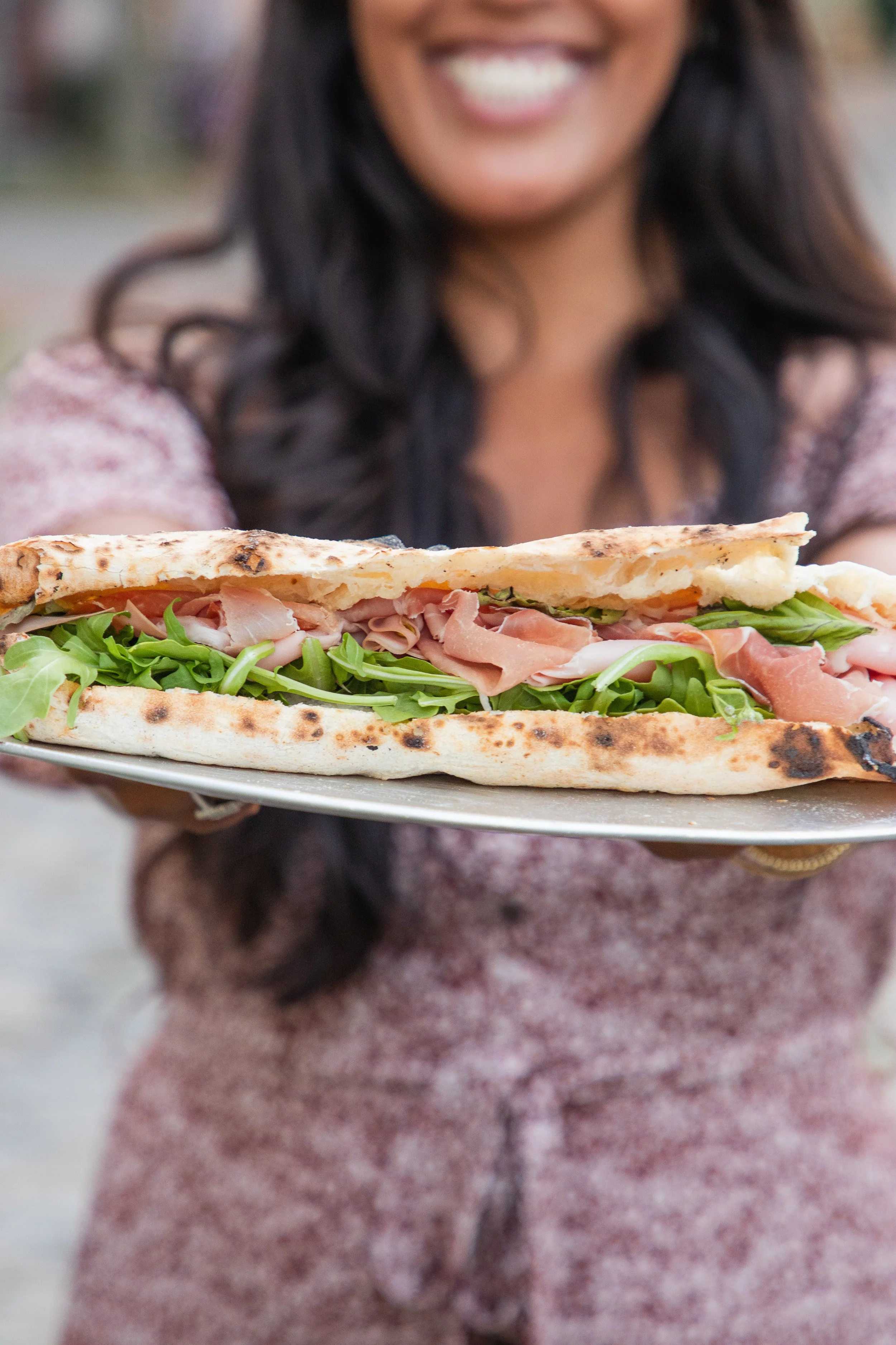 A woman holding a plate with a large sandwich with prosciutto, lettuce, and flatbread, smiling.