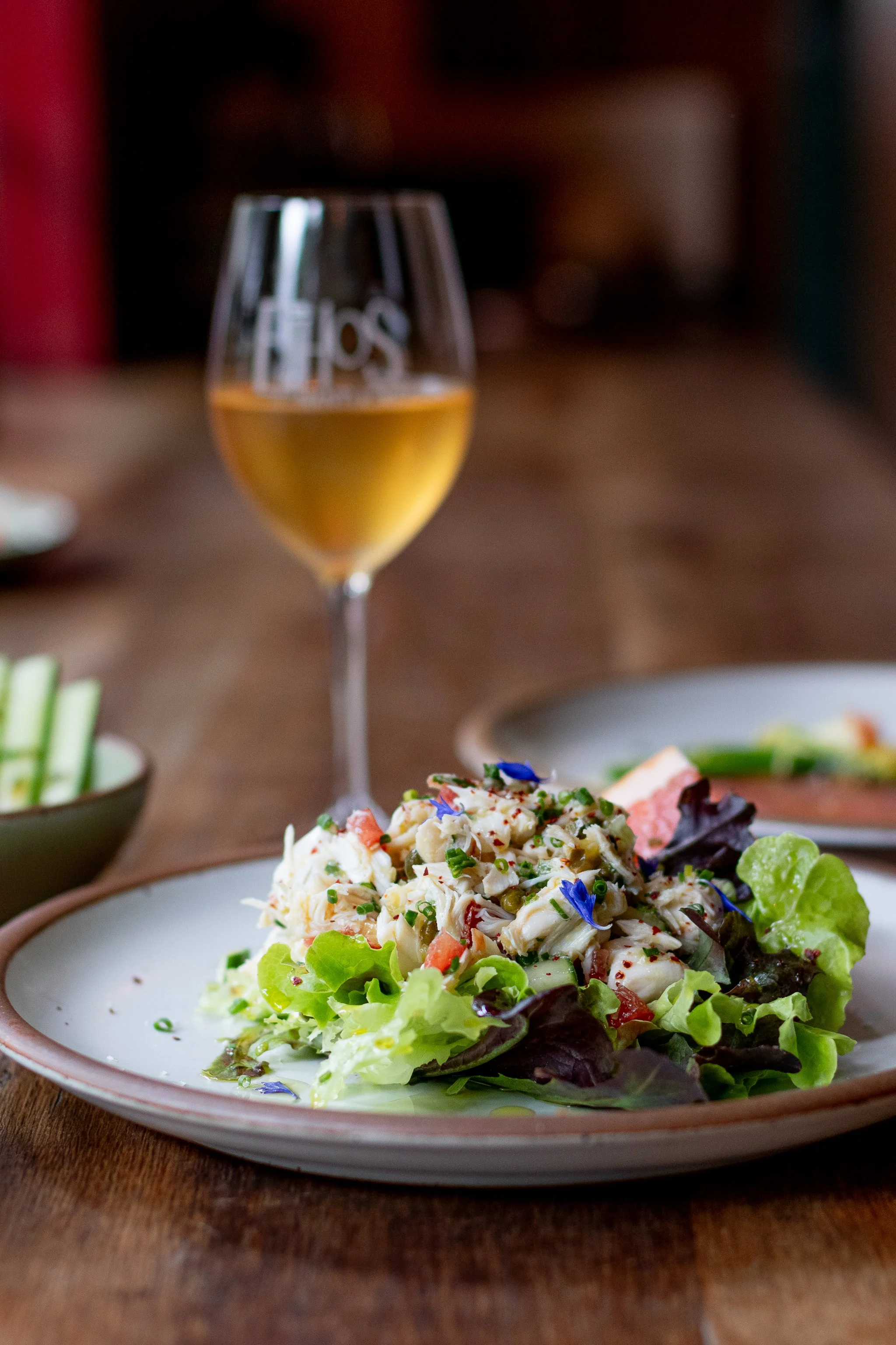 A plate of salad with lettuce, crab, and edible flowers, with a glass of white wine in the background on a wooden table.