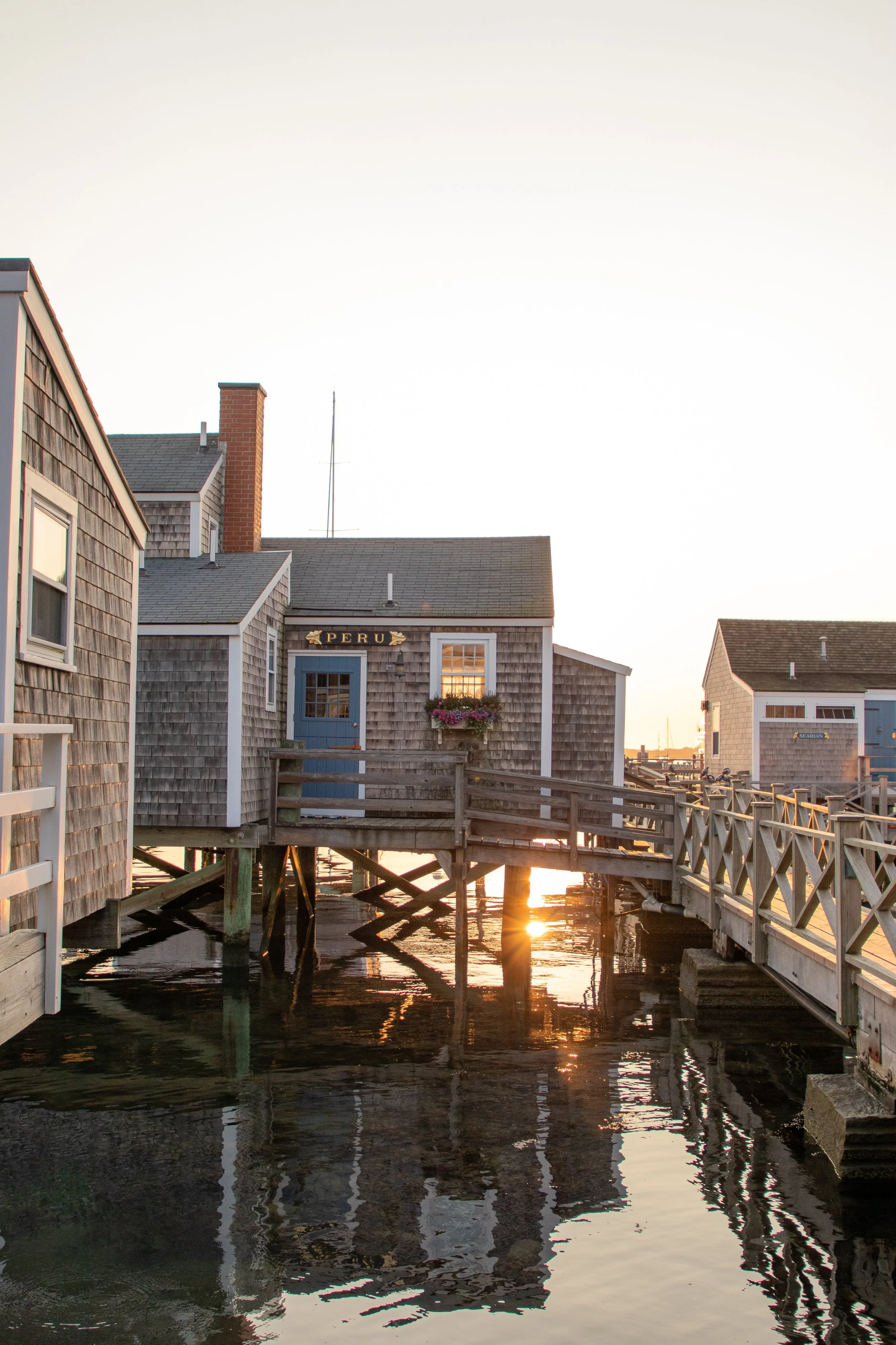 Wooden houses on stilts over water, with the sun setting behind them, including one with a sign that reads 'PERU' and a flower box on the window.
