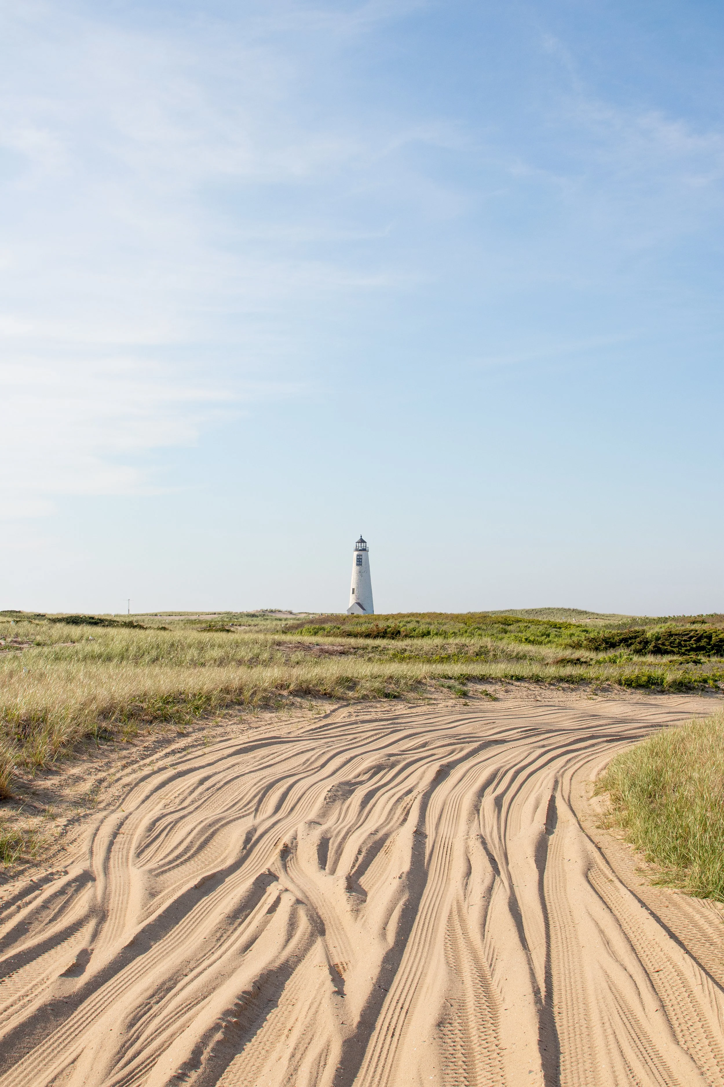 Sandy tire tracks on a beach path leading to a lighthouse in the distance, with grassy dunes and a blue sky.