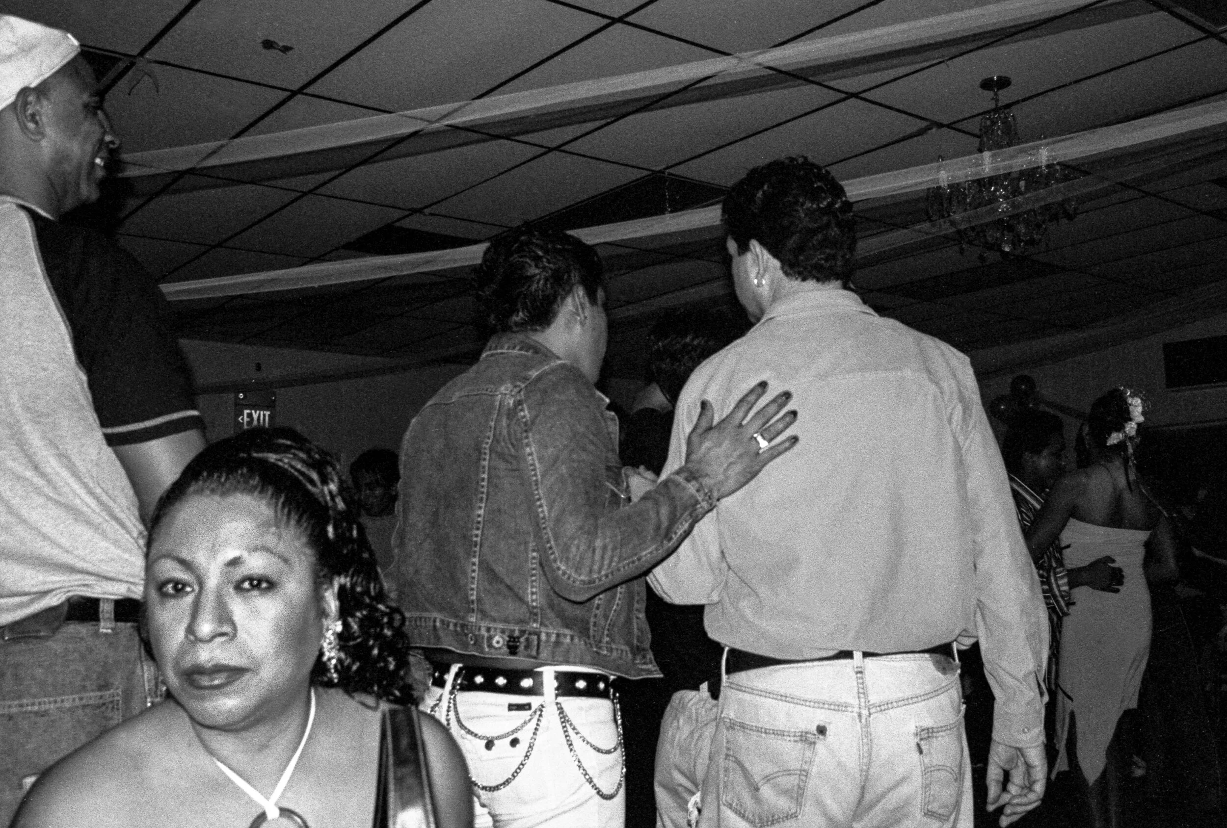 Black and white photo of people at a party, with some facing away and one woman looking at the camera, dressed in a sleeveless top with a choker necklace, in a decorated indoor setting.