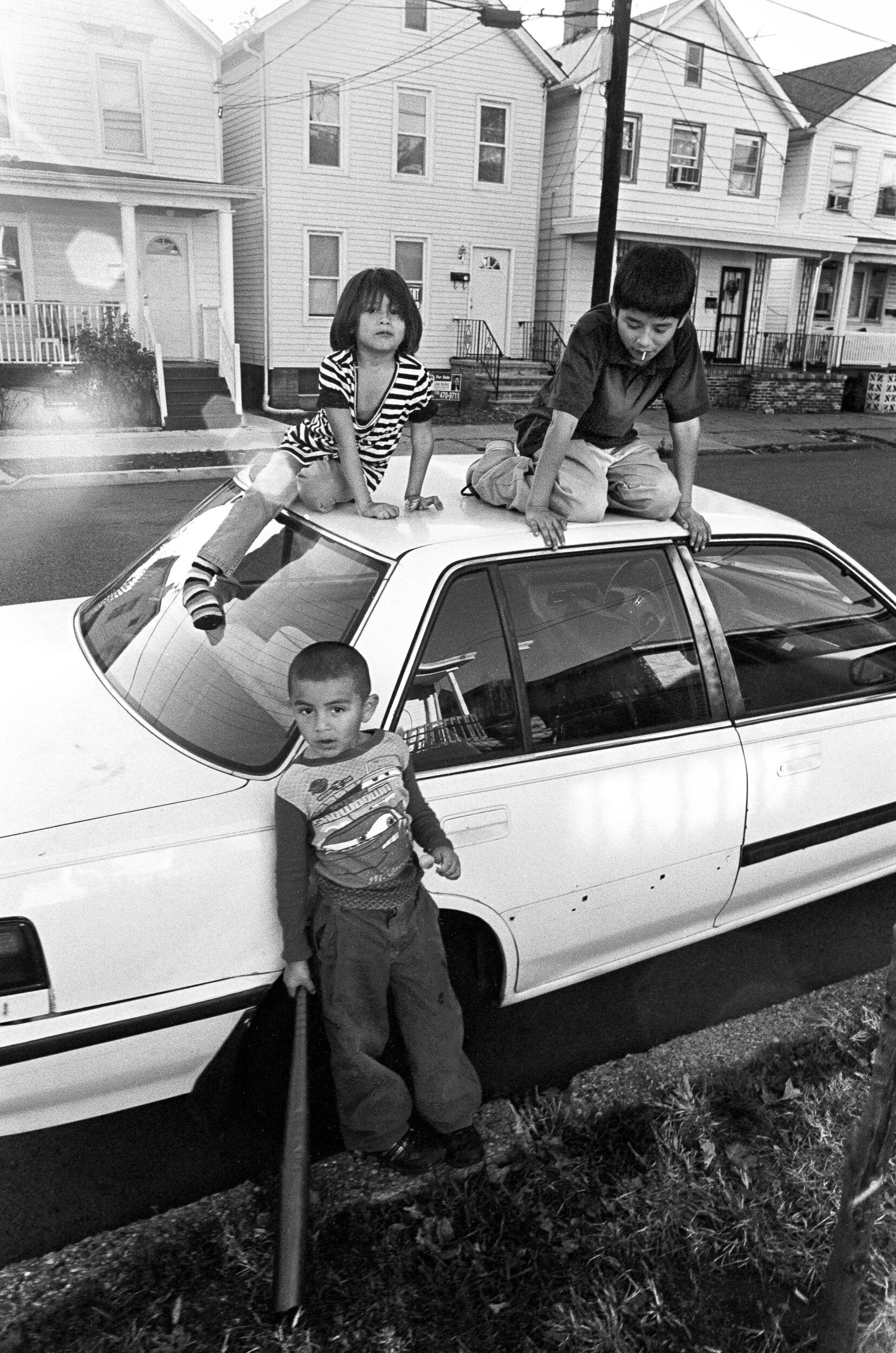 Three children playing on and around a white car parked on a residential street in front of houses, with two children on the car's roof and one standing next to it holding a long object.