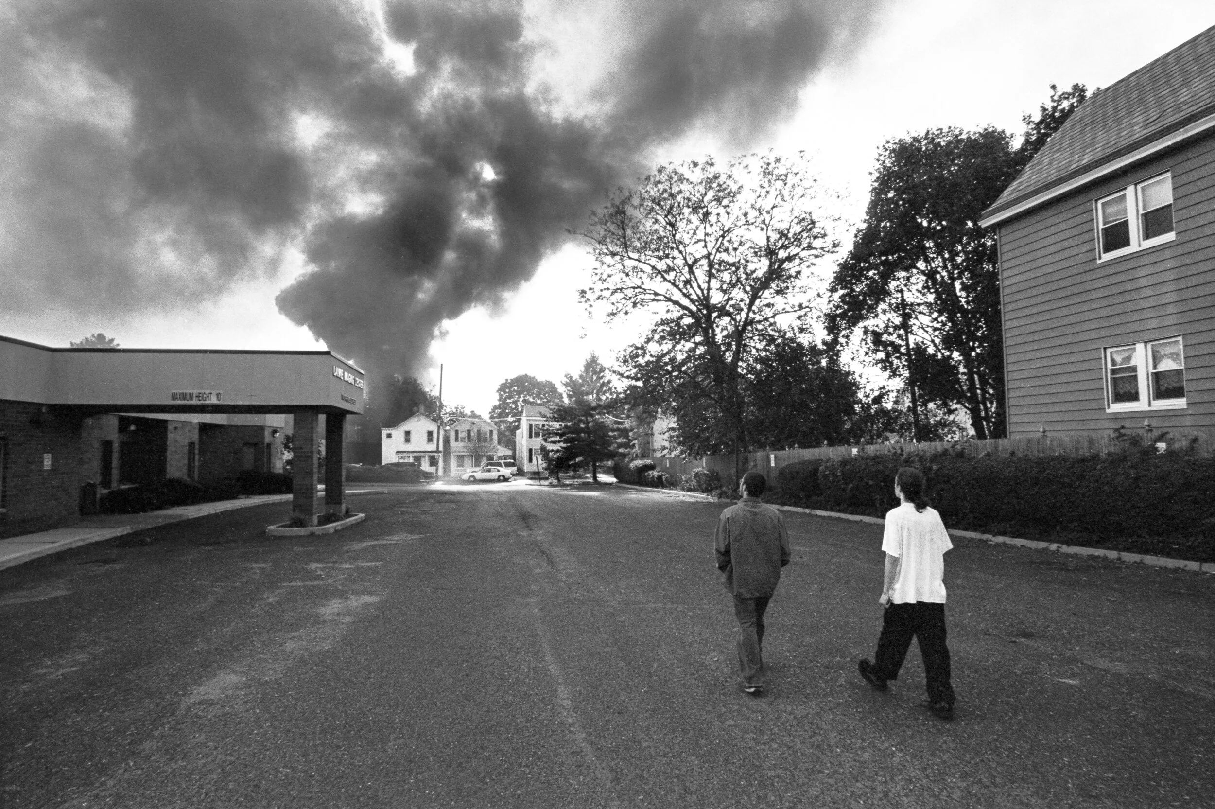 Two people walking down a street with dark smoke billowing from a fire in the background under a cloudy sky.
