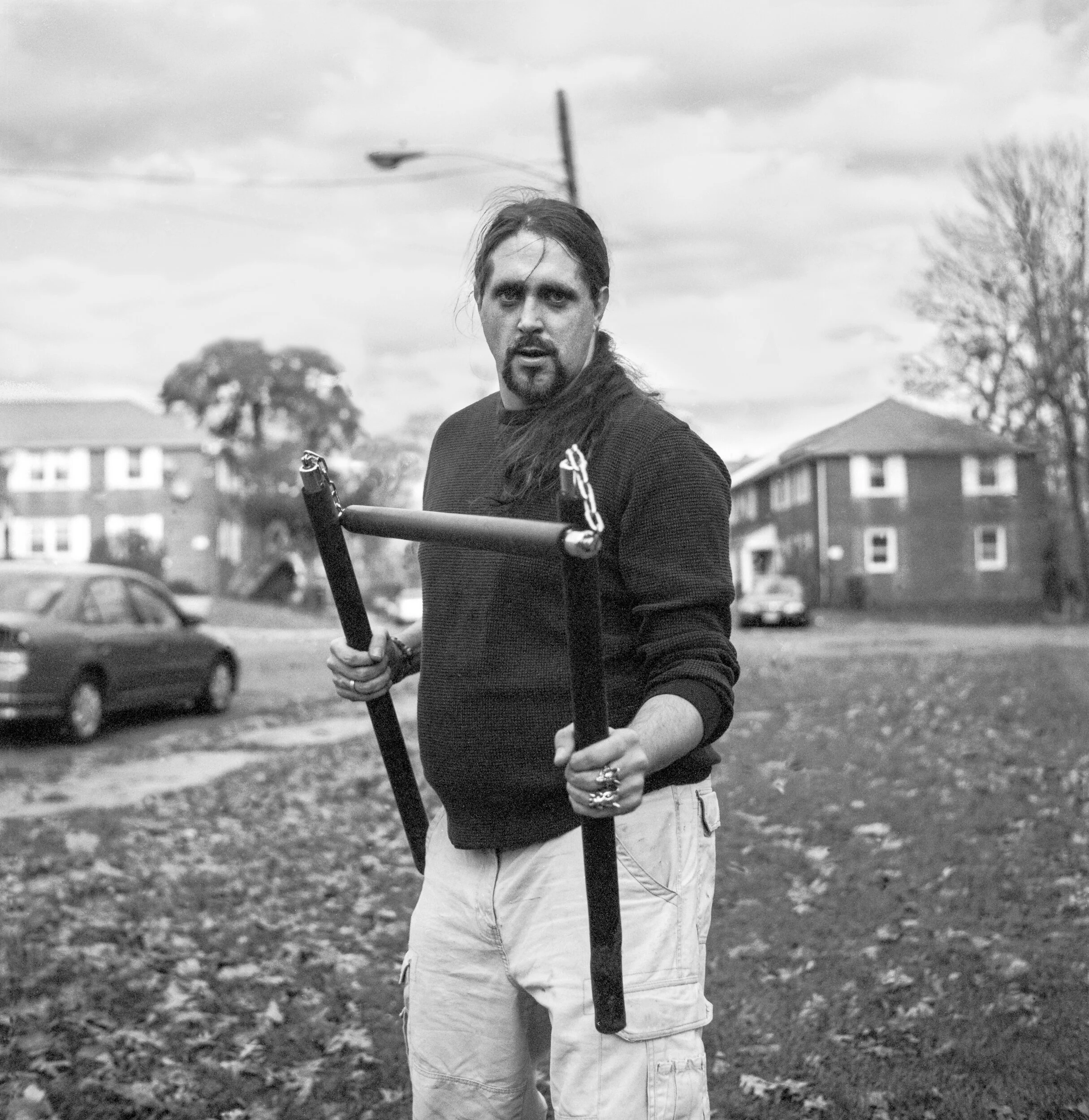 A man with long dark hair and a beard holds a tool with chains in an open area with leaves on the ground, with houses and trees in the background.
