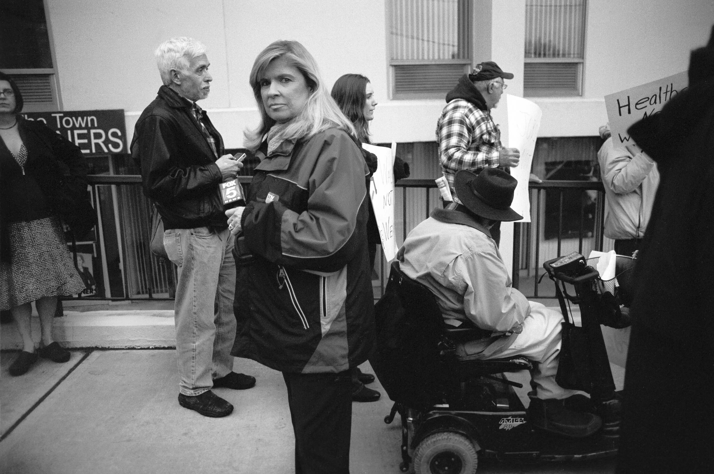 People protesting outdoors, some holding signs, including one that reads 'Health'. A woman is looking at the camera, and a man in a wheelchair is present among the crowd.