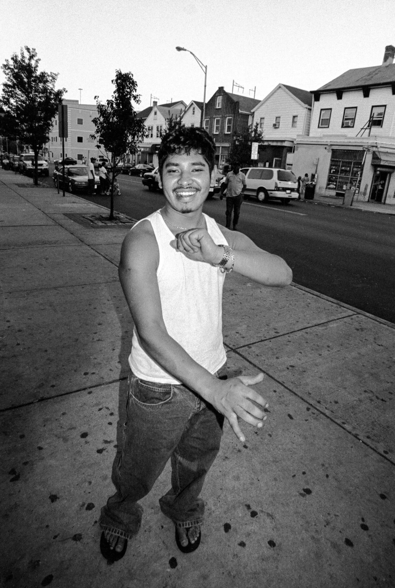 Young man standing on city sidewalk, smiling, wearing sleeveless shirt, jeans, and flip-flops, with street and buildings in the background.