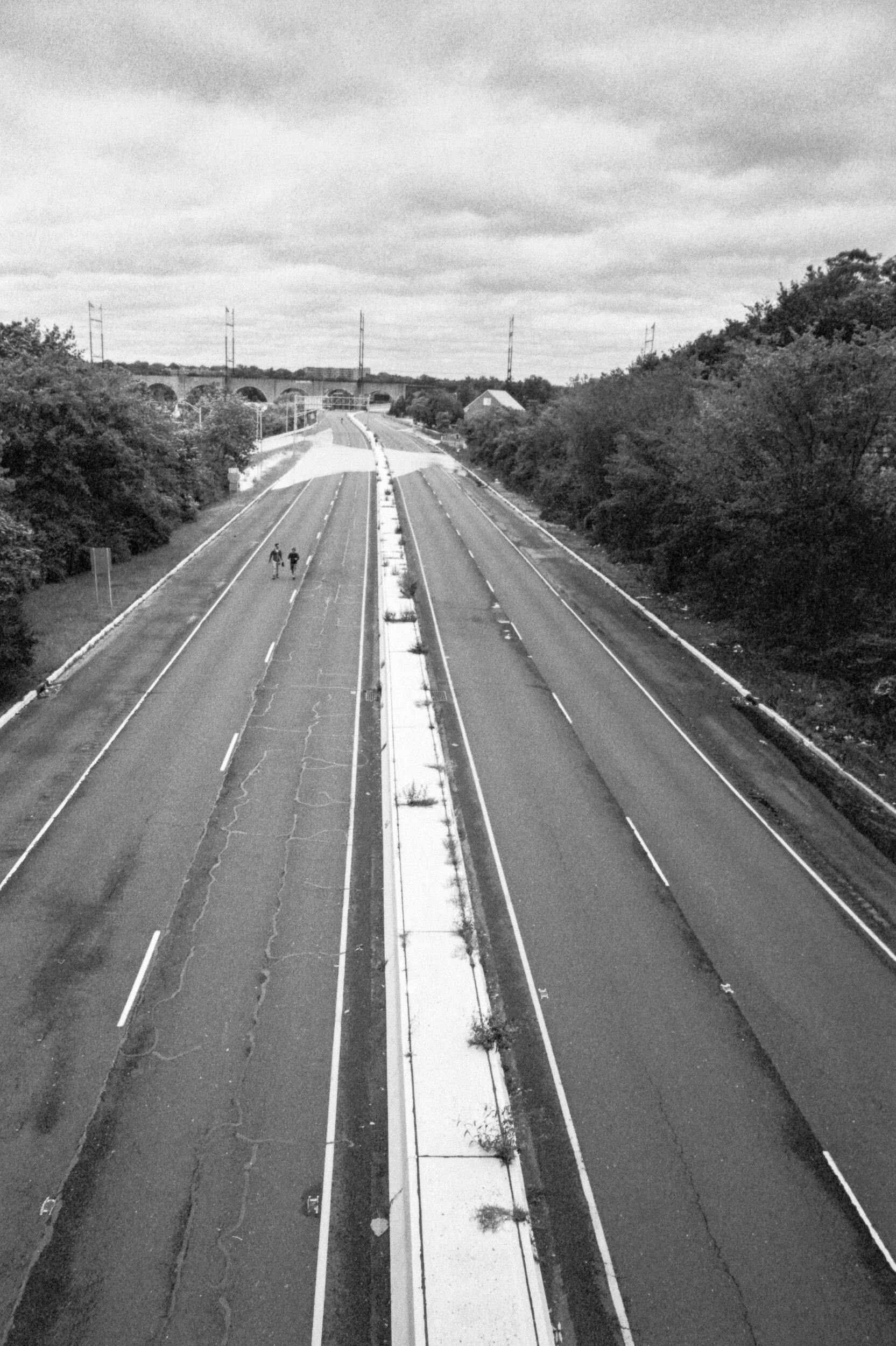 Black and white photo of an empty multi-lane road with two pedestrians walking along the middle divider, surrounded by trees on both sides under a cloudy sky.