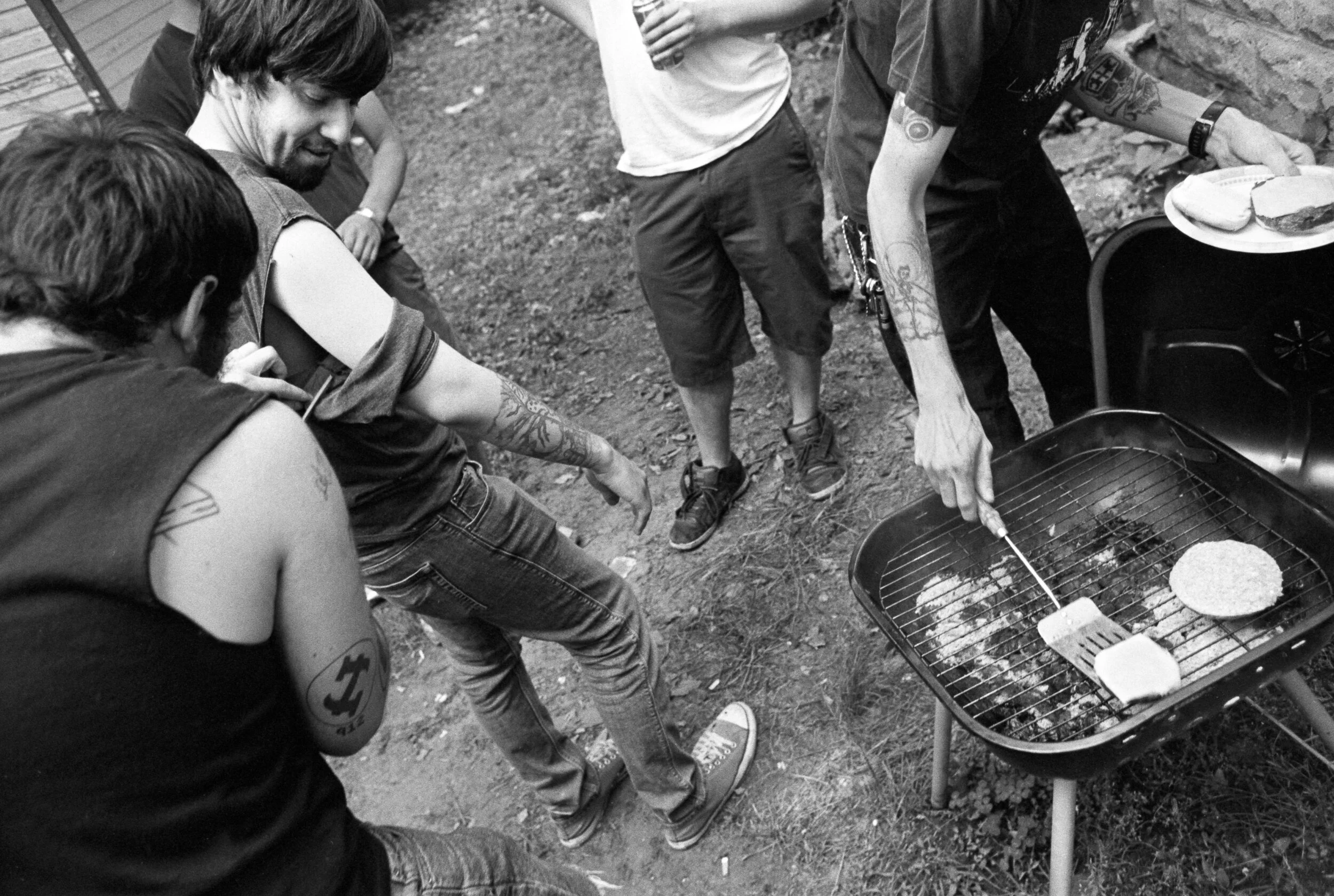 Group of people gathered around a small outdoor barbecue grill, cooking hamburgers, with one person flipping a burger.
