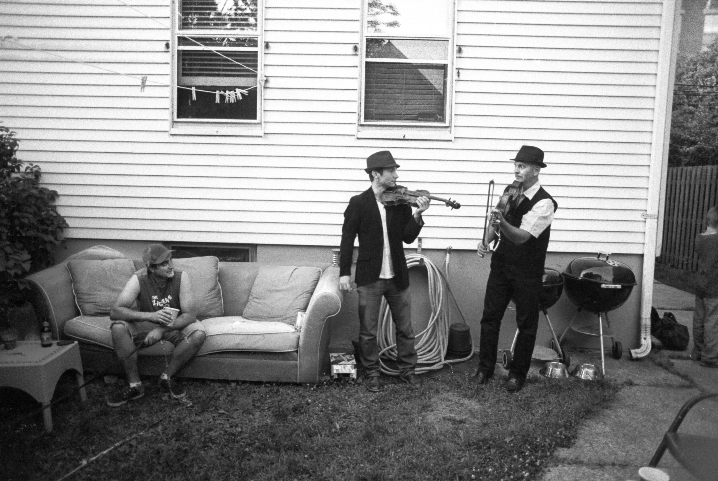 Three men are outdoors in front of a house with vinyl siding. Two men are playing guitars, one with a violin and the other with an acoustic guitar. The third man is sitting on a sofa, holding a drink, wearing a cap and sleeveless shirt. There is a gr