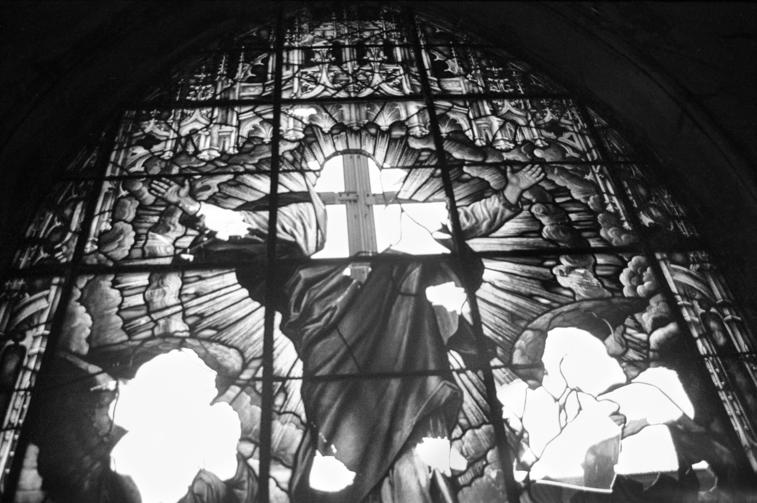 Black and white stained glass window depicting Jesus Christ with a cross, surrounded by clouds and rays of light, with some parts broken.