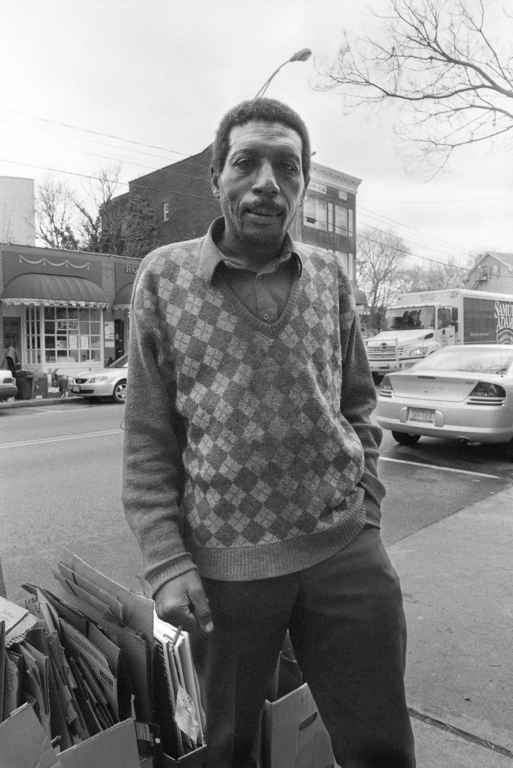A man standing on a city sidewalk near a street with parked cars and buildings in the background, captured in black and white.