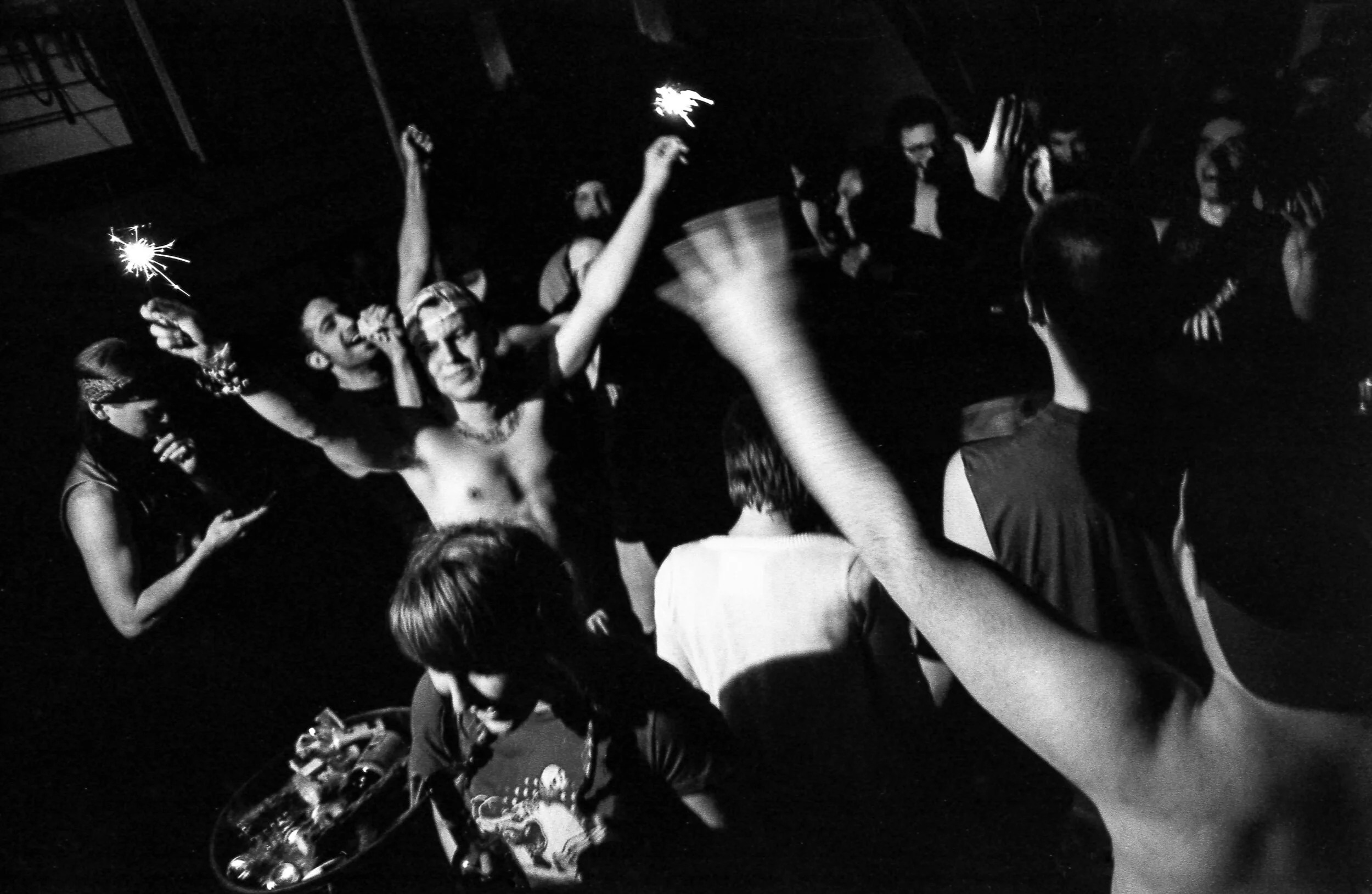 People celebrating with sparklers at a party, some dancing and others taking photos, in a dark indoor setting.
