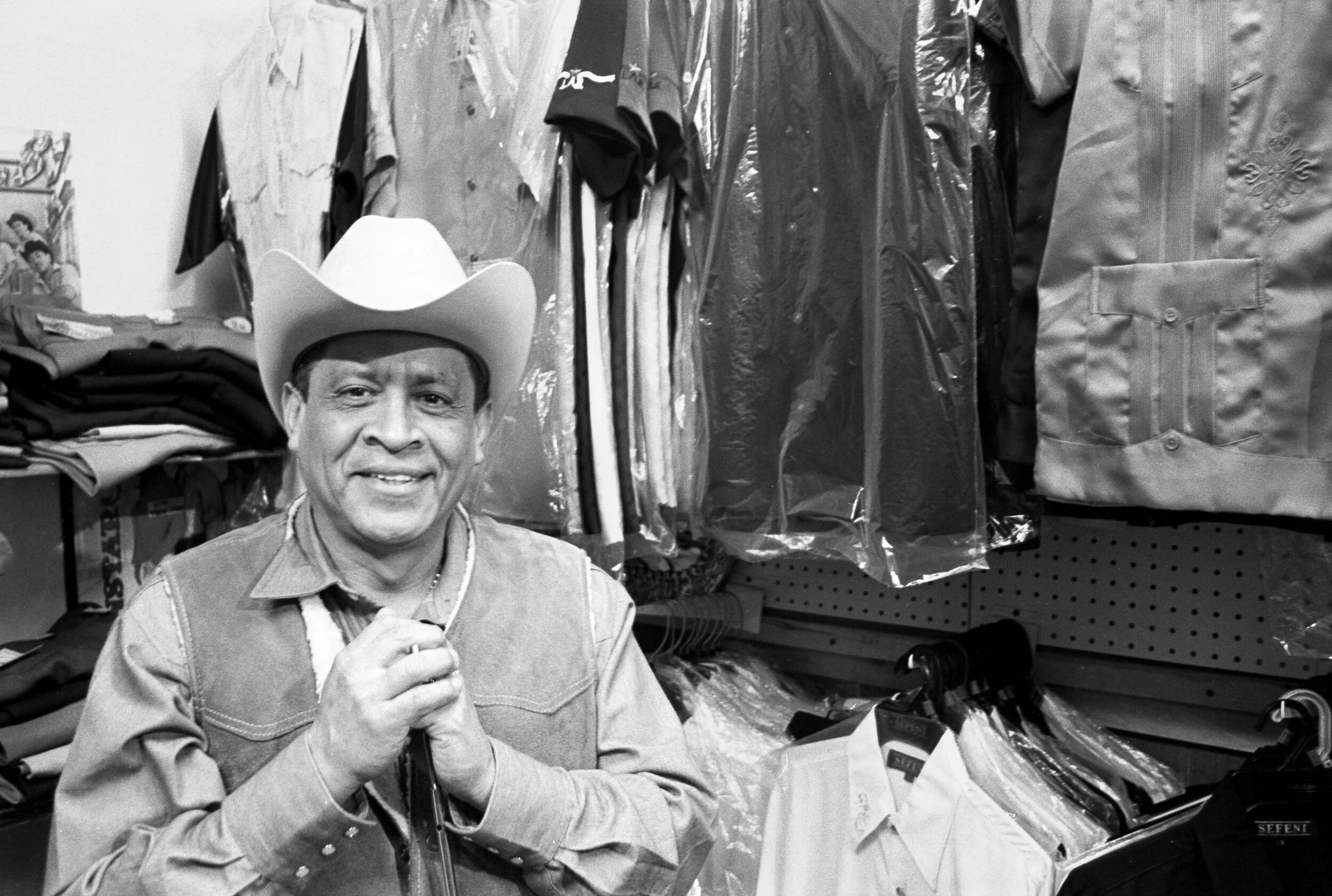 A man wearing a cowboy hat and denim vest, smiling in a clothing store with suits and shirts hanging on racks in the background.
