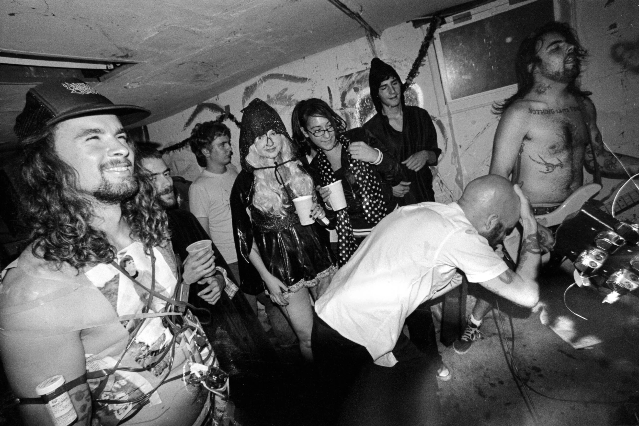 A group of people at a party in a dimly lit, graffiti-covered basement. Some are holding drinks, and one person is DJing with turntables while others watch and enjoy the scene.