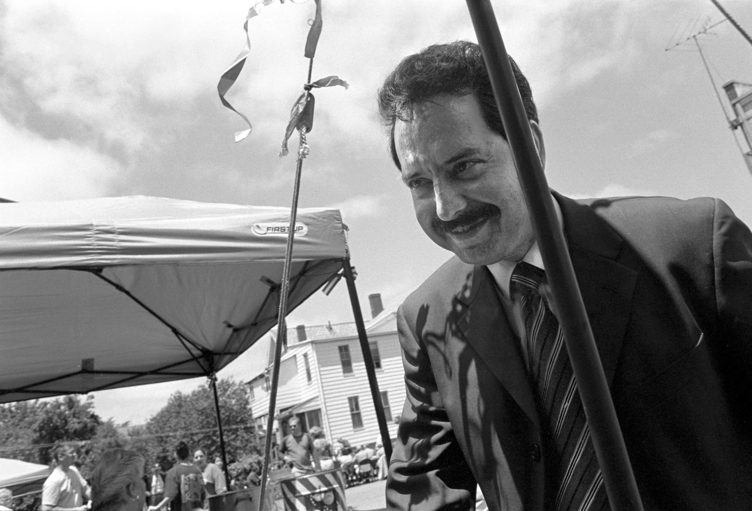 A black-and-white photo of a man in a suit and striped tie smiling at an outdoor event, with tents and people in the background.