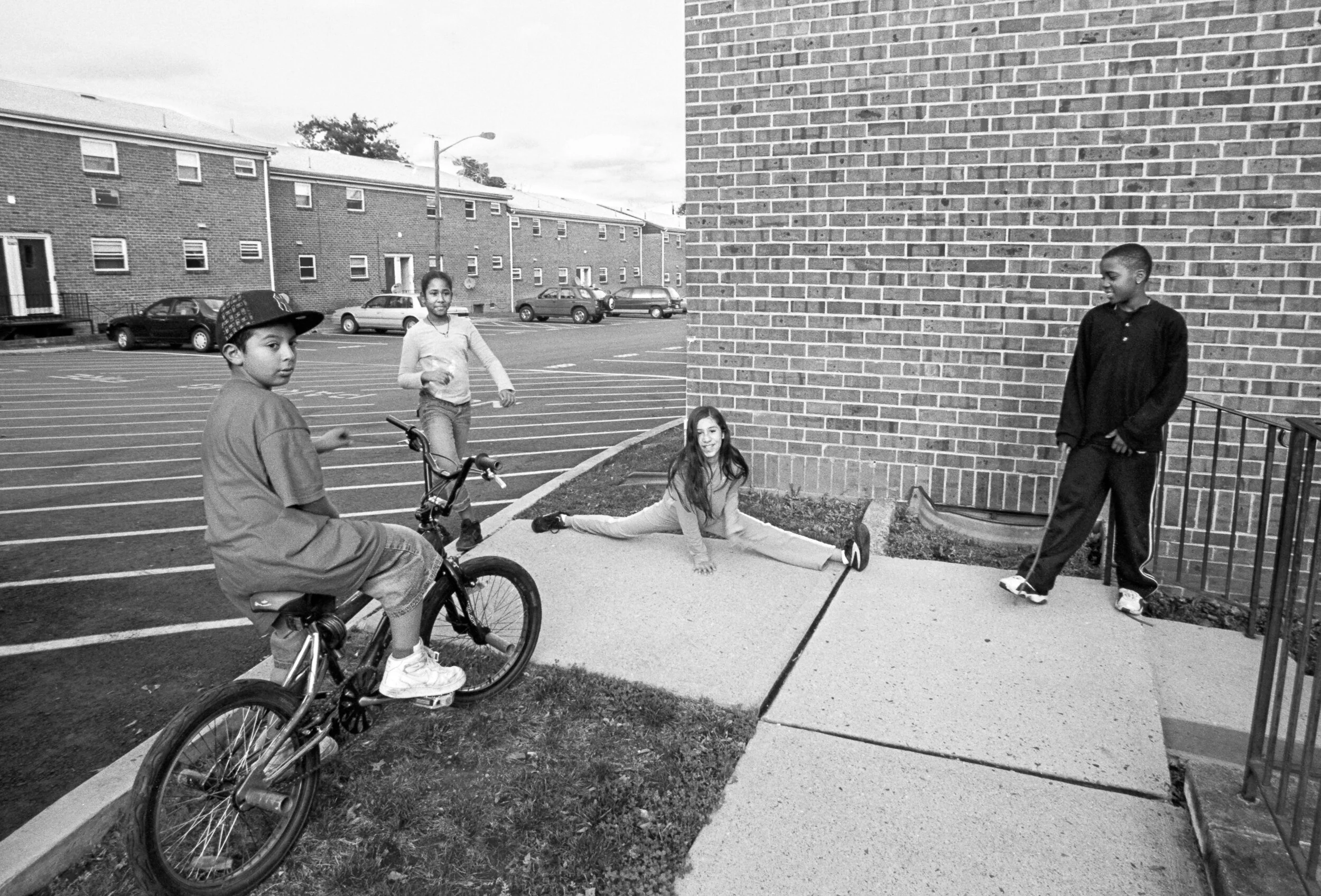 Four children playing outside near a brick building, with a parking lot in the background; one girl is doing splits on the sidewalk while a boy on a bicycle and two other children stand nearby.