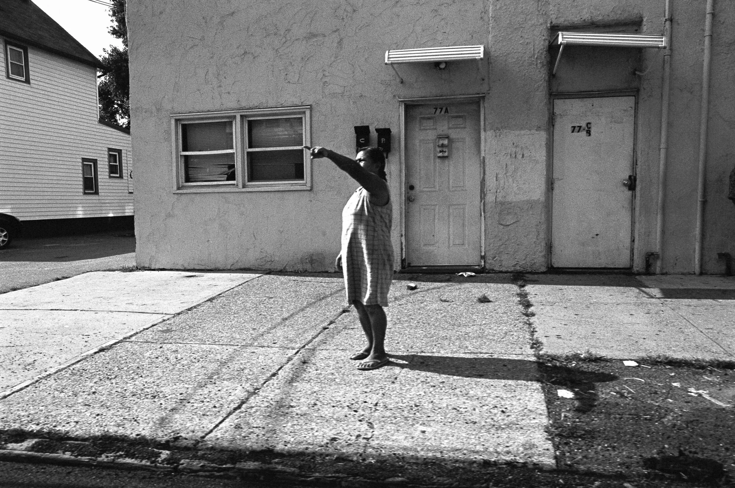 A man standing on a sidewalk, pointing to the left, outside a building with two doors and two windows, in black and white.