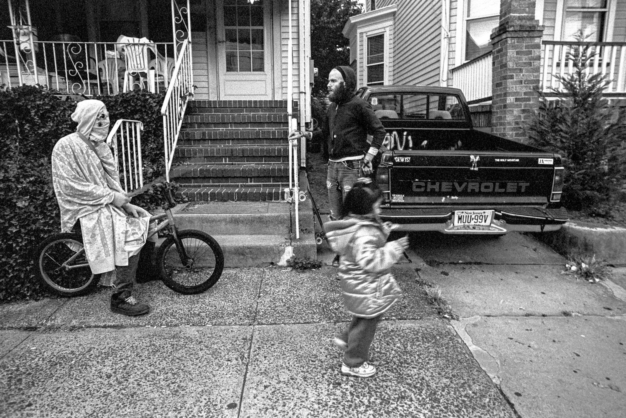 A black and white photo of a woman sitting on a bicycle, a young girl walking by, and a man standing by a Chevrolet pickup truck on a residential street with stairs leading to a house.