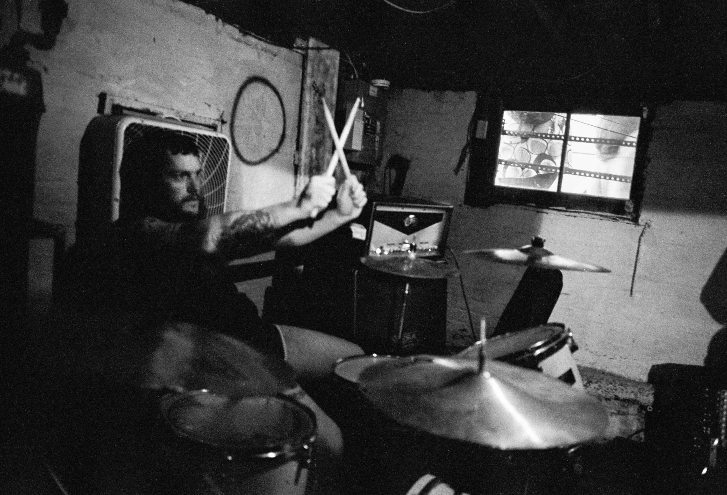 A man with tattoos playing drums in a dimly lit room, with a television showing a video and a fan on the wall behind him.