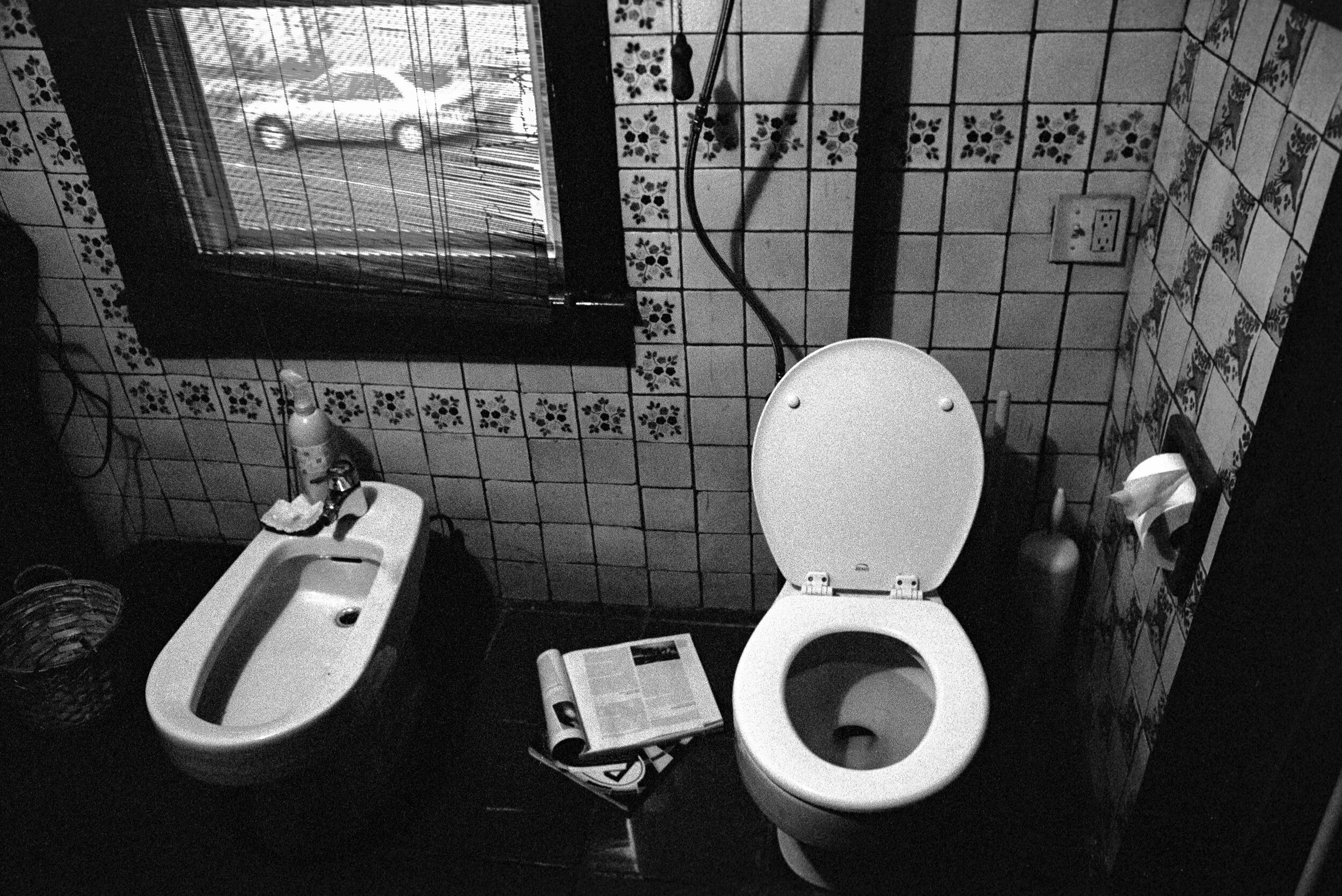 A black and white photo of a bathroom with a toilet, a bidet, a window with blinds, a mirror, a soap bottle, a roll of toilet paper, and a partially open newspaper on the floor.