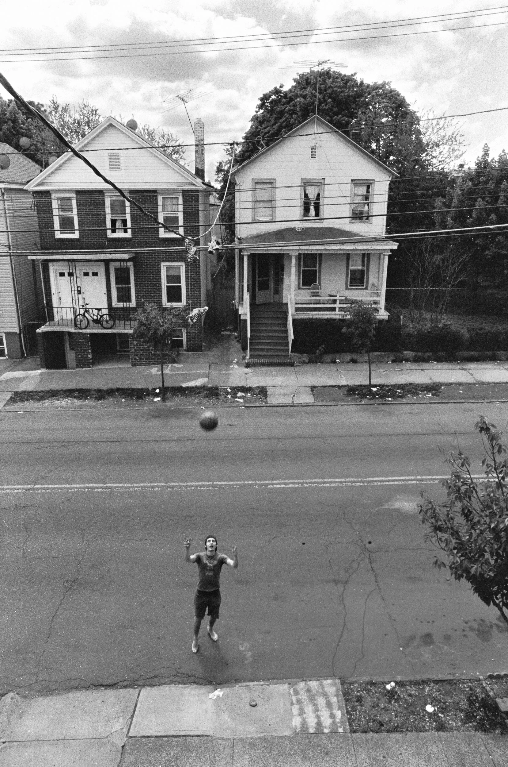 A man playing basketball outside on a street, seen from above. The photo is in black and white, showing street houses with sidewalks and power lines overhead.