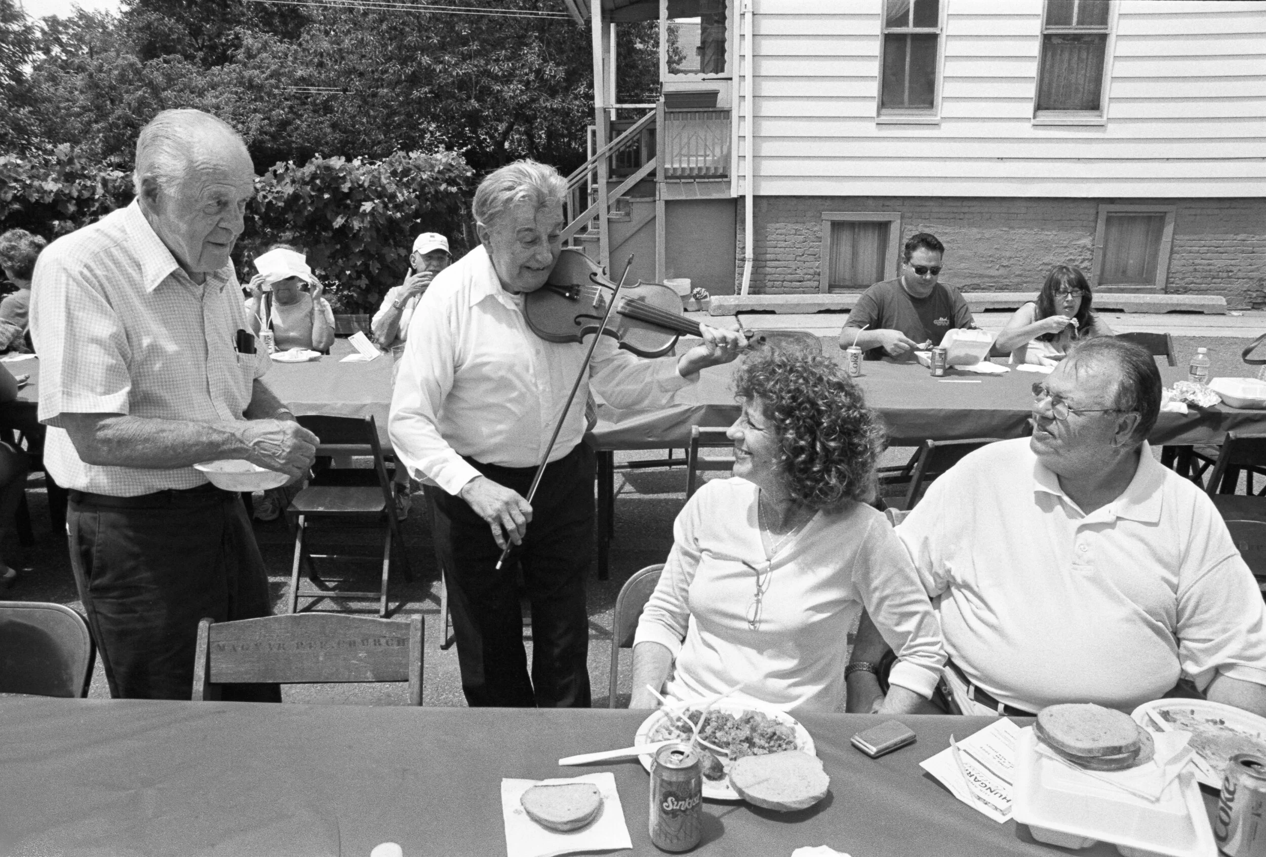 Black and white photo of people at an outdoor gathering. Two elderly men are standing, one is holding a bowl and the other is playing a violin. Two women are seated at a table, smiling and listening. The table has food and drinks on it, with plates, 