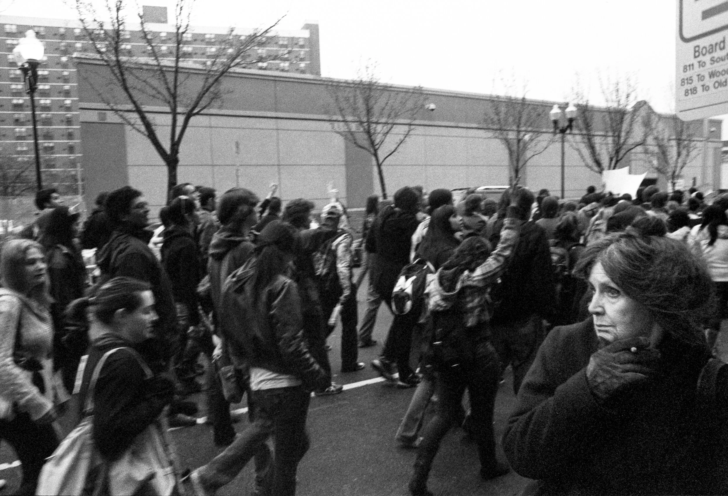Black and white photo of a large group of women marching on a city street, some raising their fists, with leafless trees and buildings in the background.