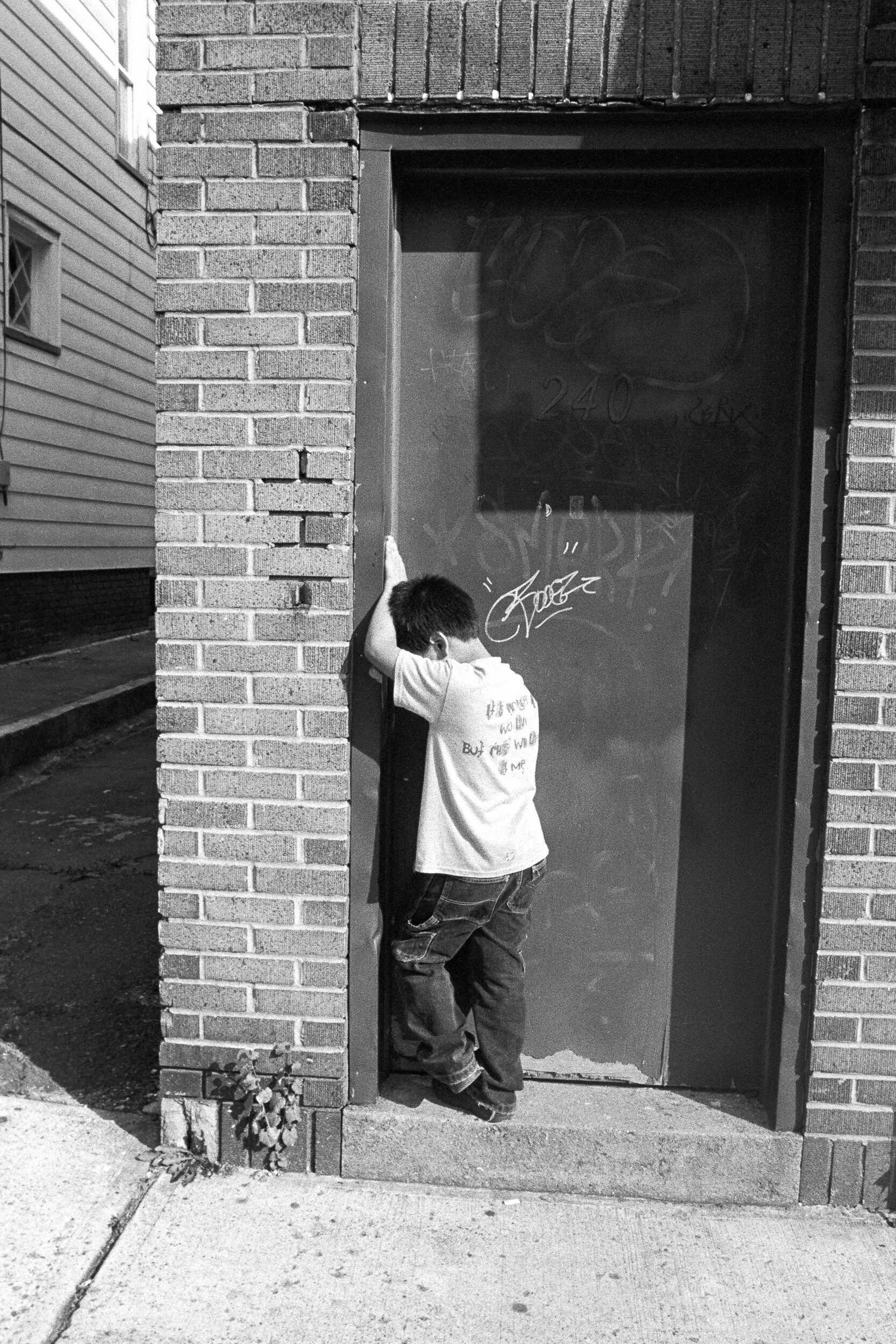 A child leaning against a brick wall and a door with graffiti, on a sidewalk in a residential area, black and white photo.