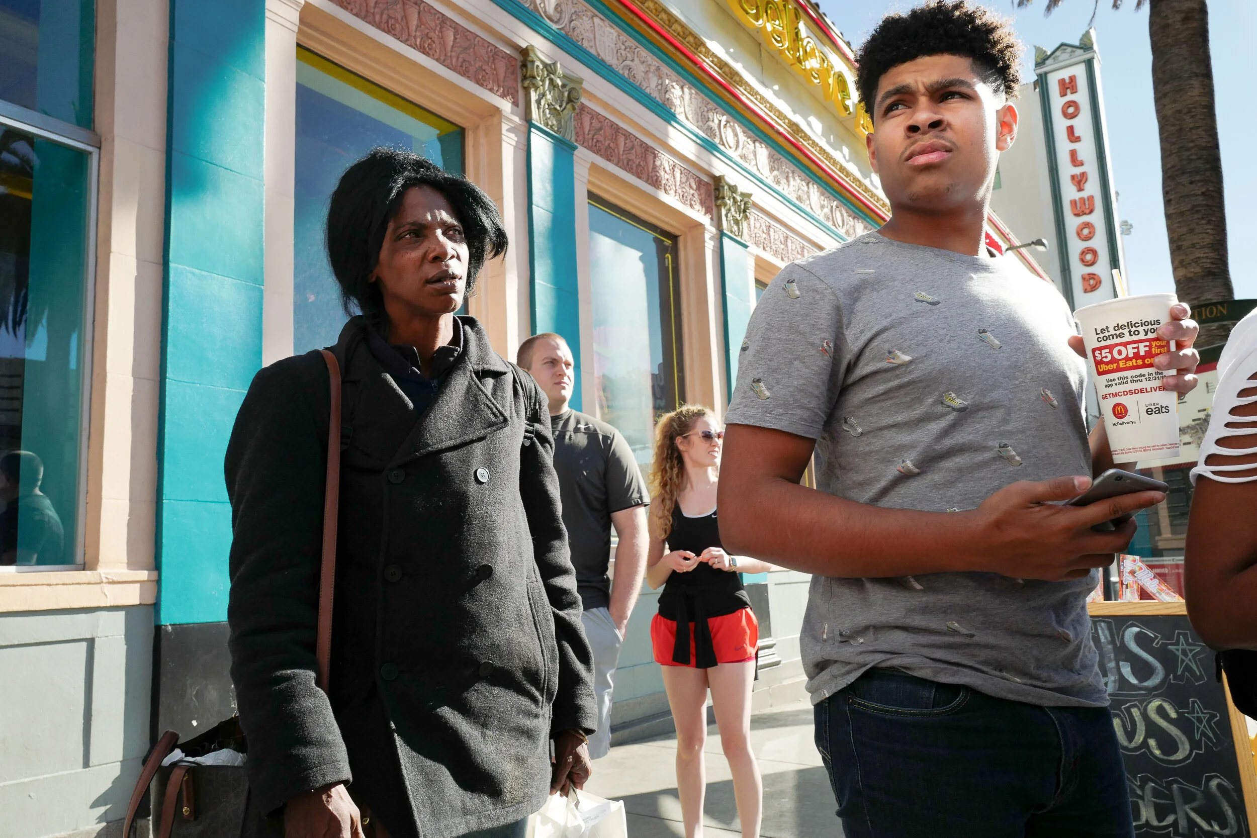 People standing on a city sidewalk in front of a colorful building with a sign that reads 'Hollywoo'. A young man is holding a cup and a phone, while a woman looks on with a concerned expression.