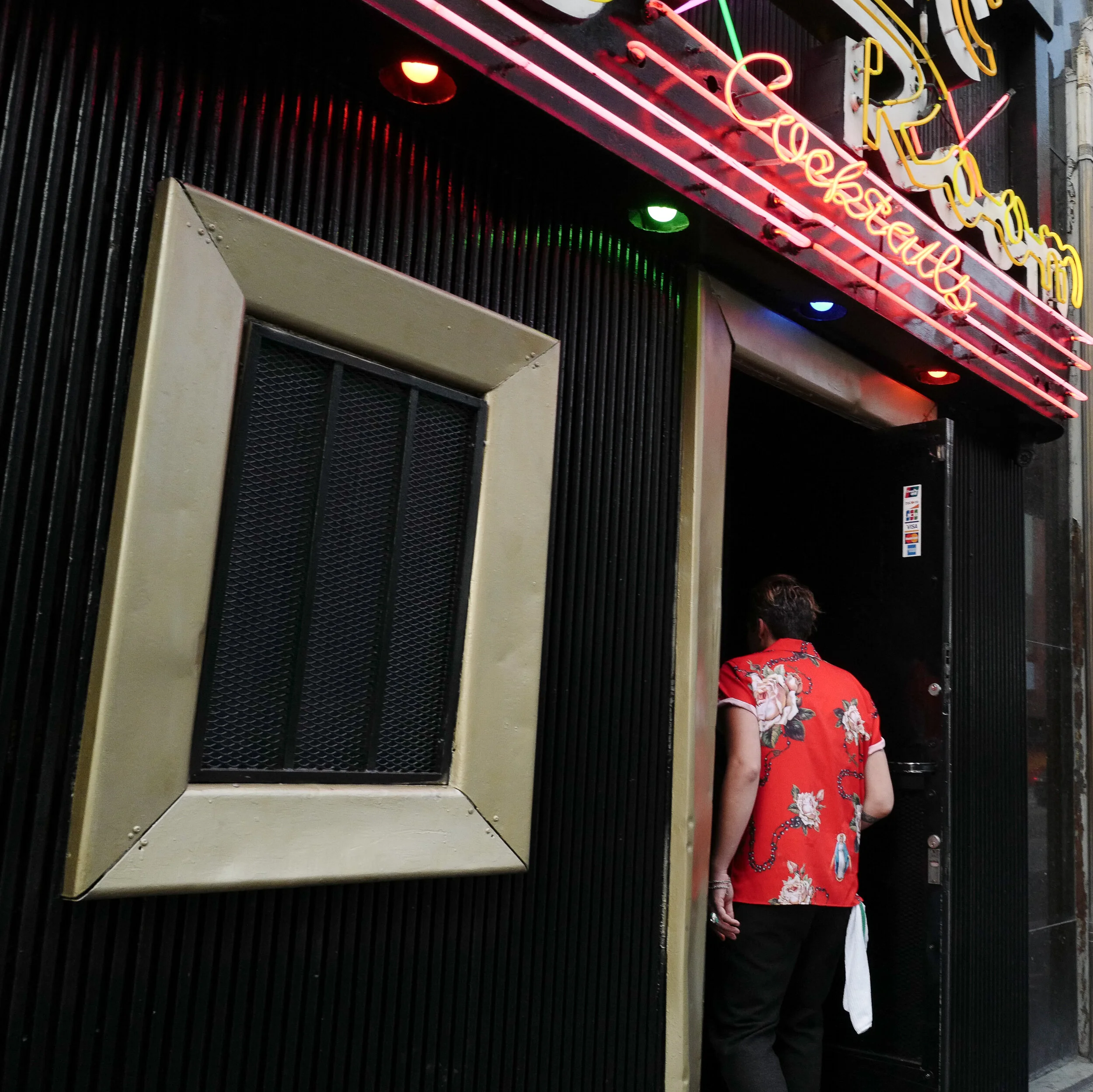 Person wearing a red floral shirt entering a nightclub with a neon sign that says 'Tequila' in bright colors
