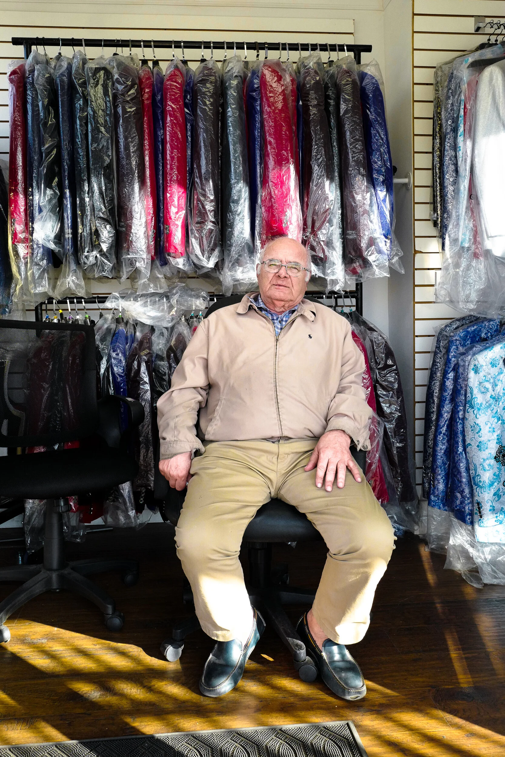 An older man with glasses, wearing a beige jacket and khaki pants, sitting in an office chair inside a dry cleaning store, with racks of plastic-covered suits and dresses in the background.