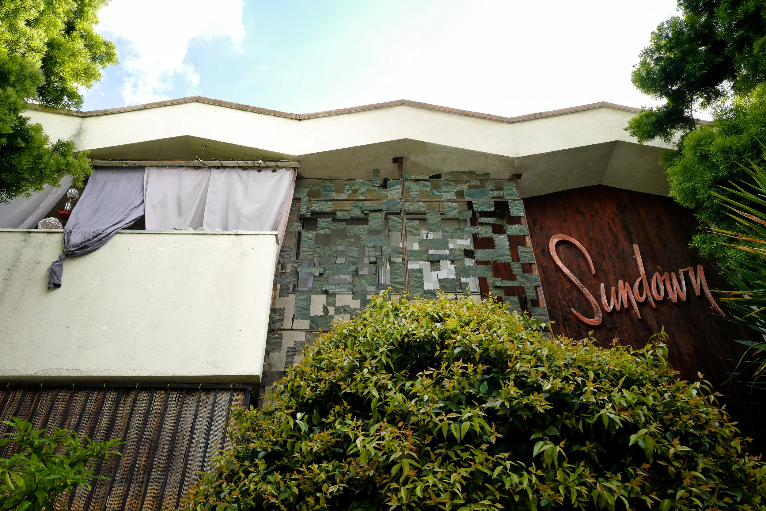 The facade of a building named "SunDour" with a decorative wall, a balcony with hanging laundry, and lush greenery in front.