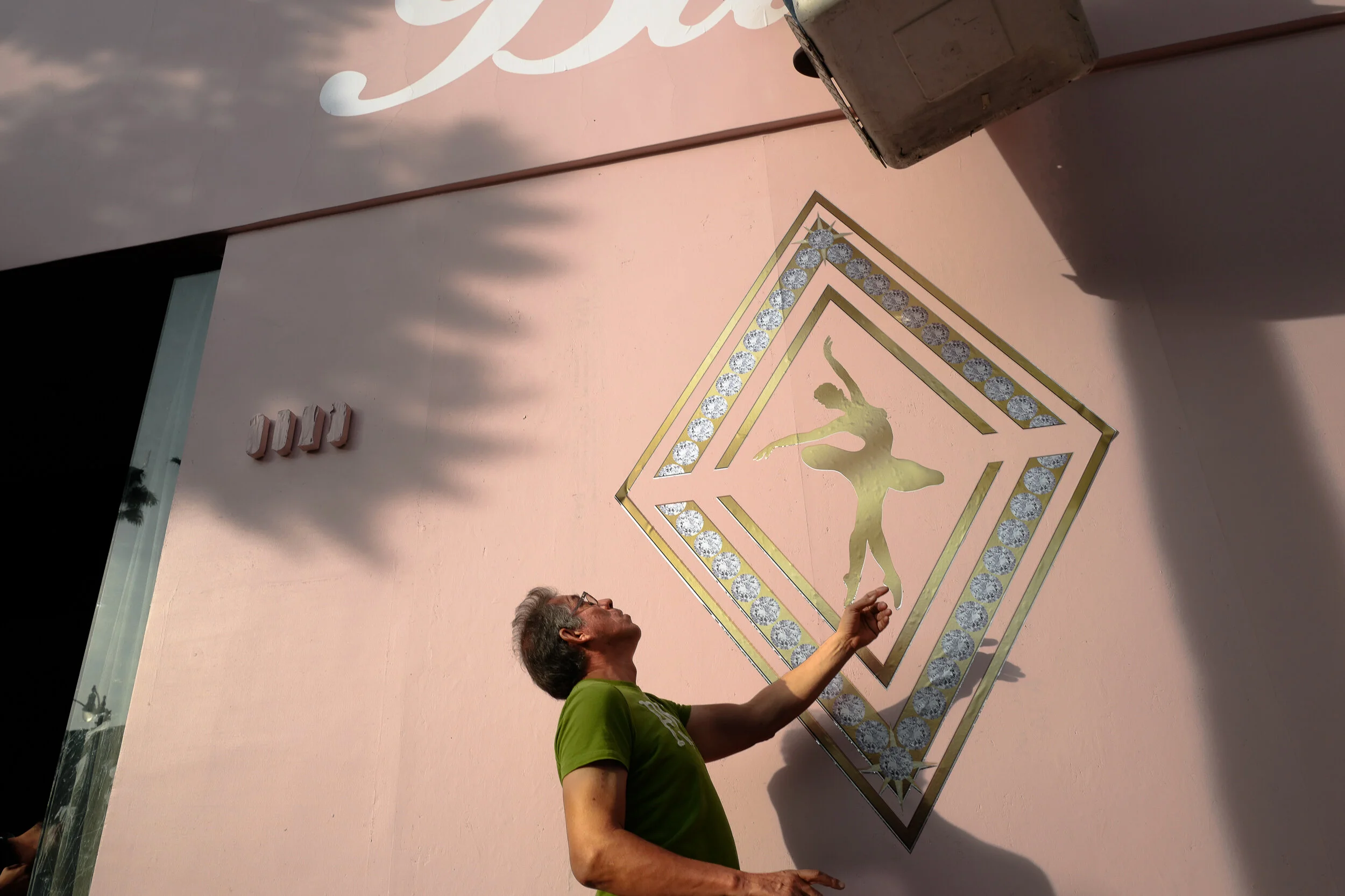 Man in green shirt standing in front of a pink wall with dance-themed mural and decorative frame, partly in shadow, with a large metallic container nearby.