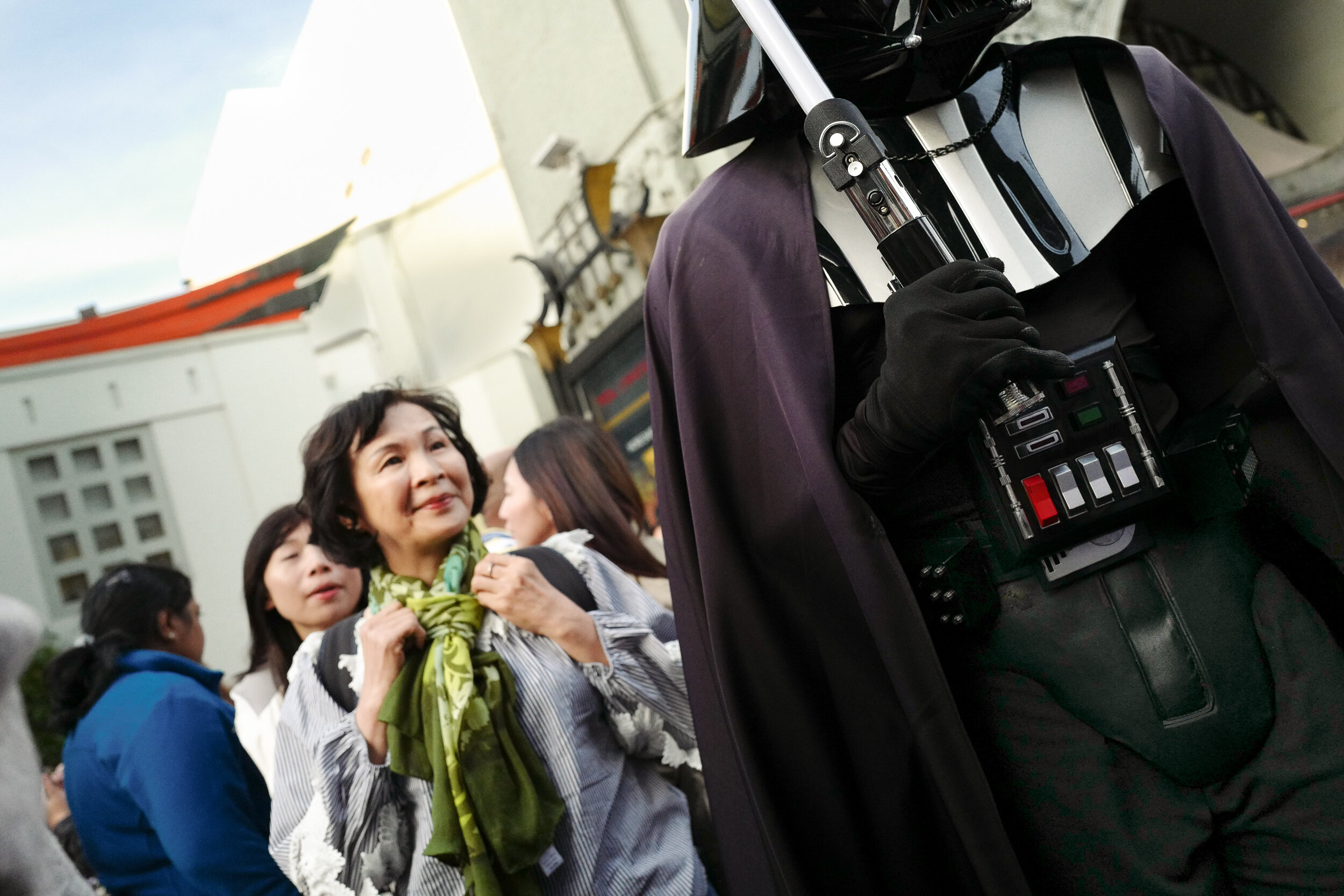 Group of people, mostly women, standing outdoors, watching a person in a Darth Vader costume from Star Wars, who is holding a remote control device.