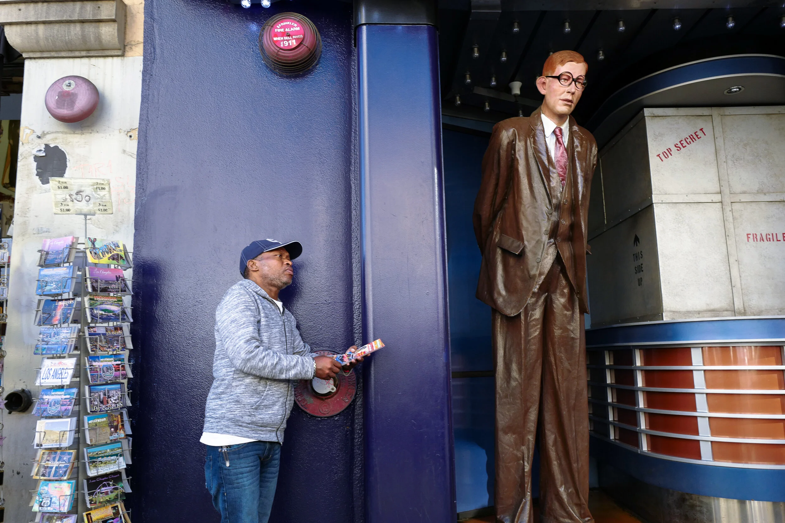 A man stands on the sidewalk near a large figure of a man in a brown suit. The figure is a statue or sculpture. There are postcards and a flyer display on the left side of the image. The scene appears to be outside a building or attraction.