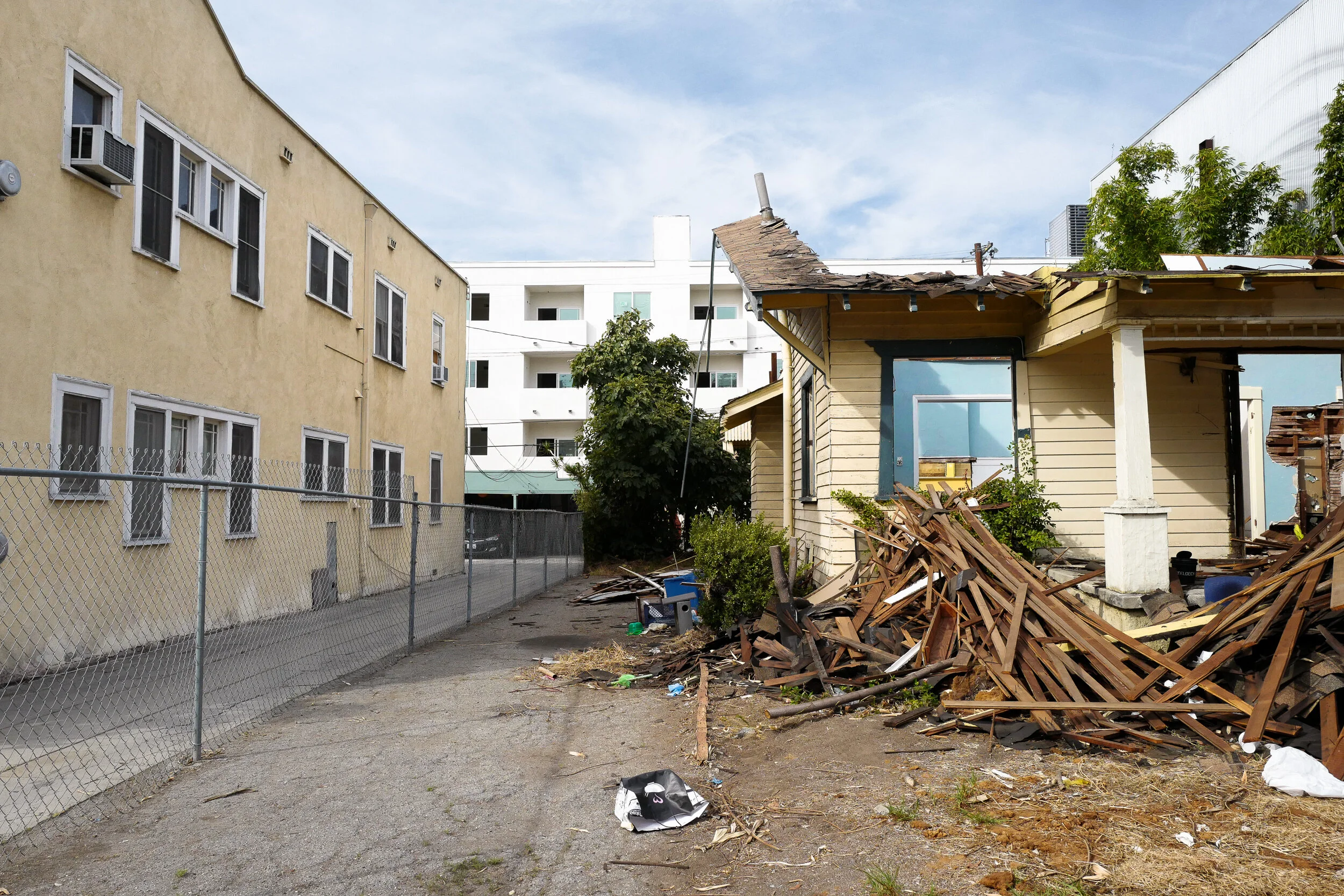 Backyard with damaged yellow house, pile of wooden debris, and neighboring apartment buildings under a cloudy sky.