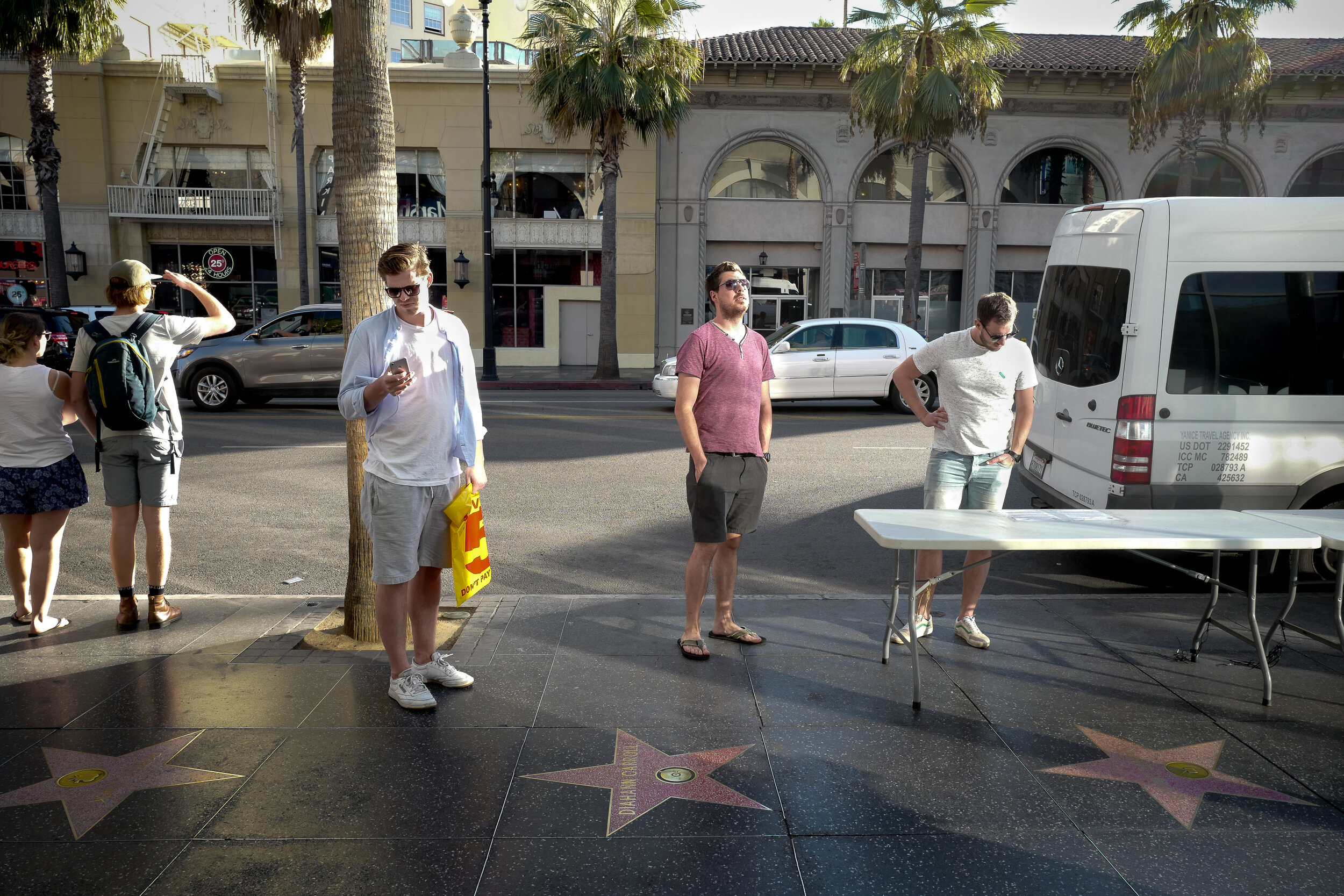 People standing on Hollywood Walk of Fame sidewalk with stars embedded in pavement, some looking at their phones, in front of palm trees and city buildings.