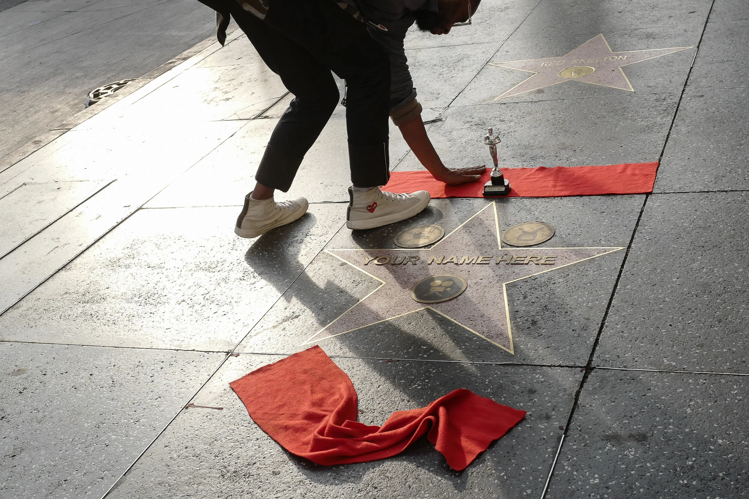 Person placing a trophy on a star on the Hollywood Walk of Fame with space for a custom name, red cloth on the sidewalk, other stars visible, and a star labeled 'RECORDING.'