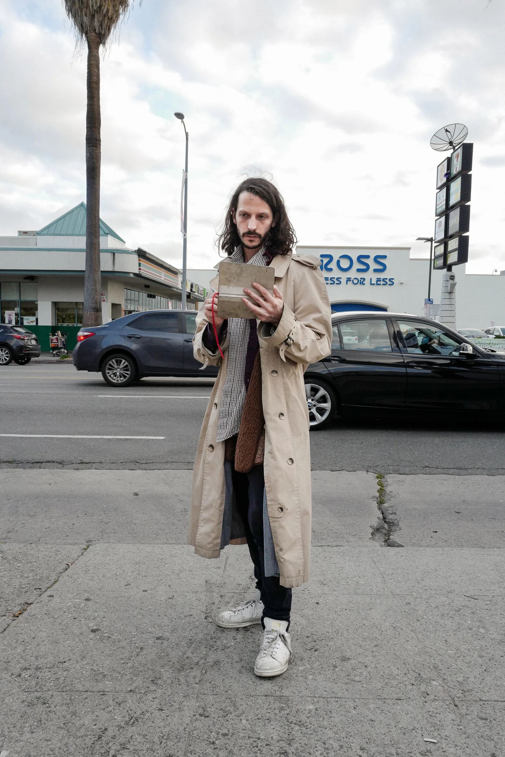 A man with long dark hair and a beard, wearing a beige trench coat and white sneakers, stands on the sidewalk looking at a tablet device. Behind him, there is a street scene with cars and a large retail store with the sign 'ROSS'.