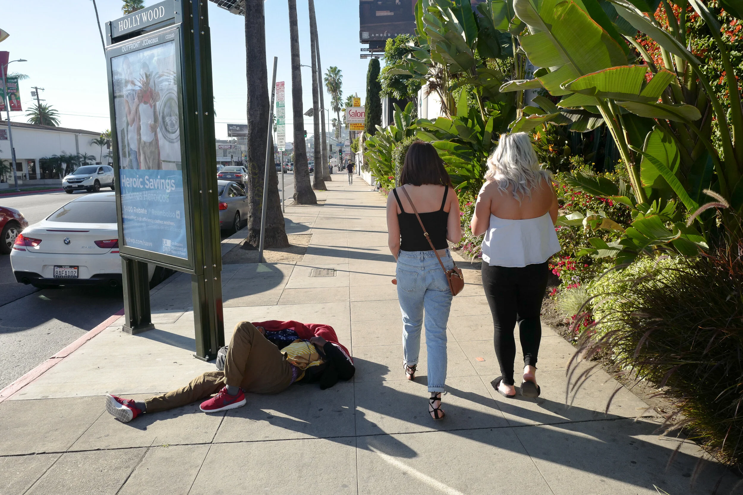 Two women walking on a sidewalk with lush greenery on the right side and parked cars on the left, while a man is lying on the ground nearby.
