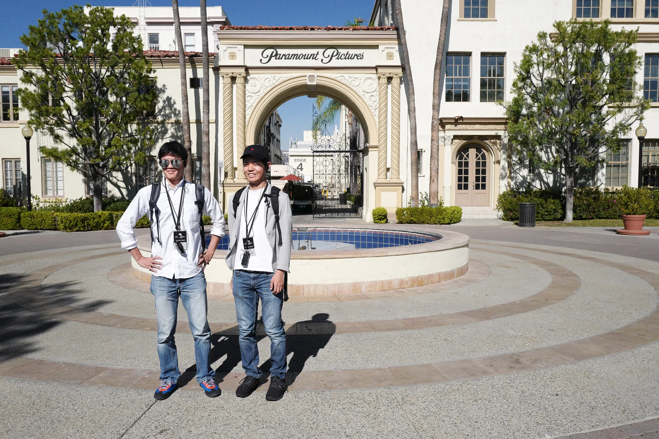 Two young men standing in front of the Paramount Pictures sign at the entrance to the studio lot. They are wearing casual clothes, backpacks, and authorization badges, smiling at the camera on a sunny day.